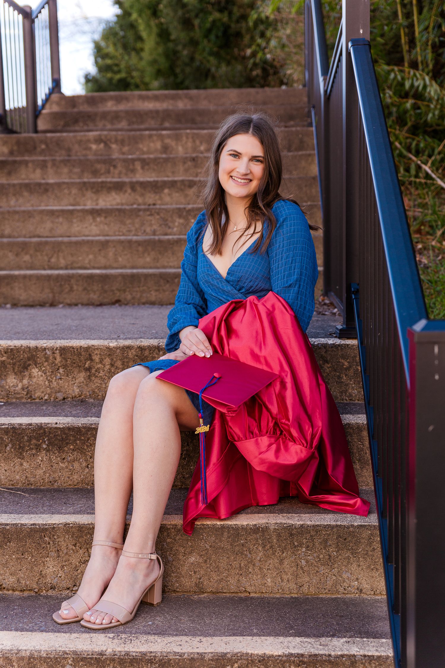 Graduate in blue sweater sits on concrete steps with red graduation gown.