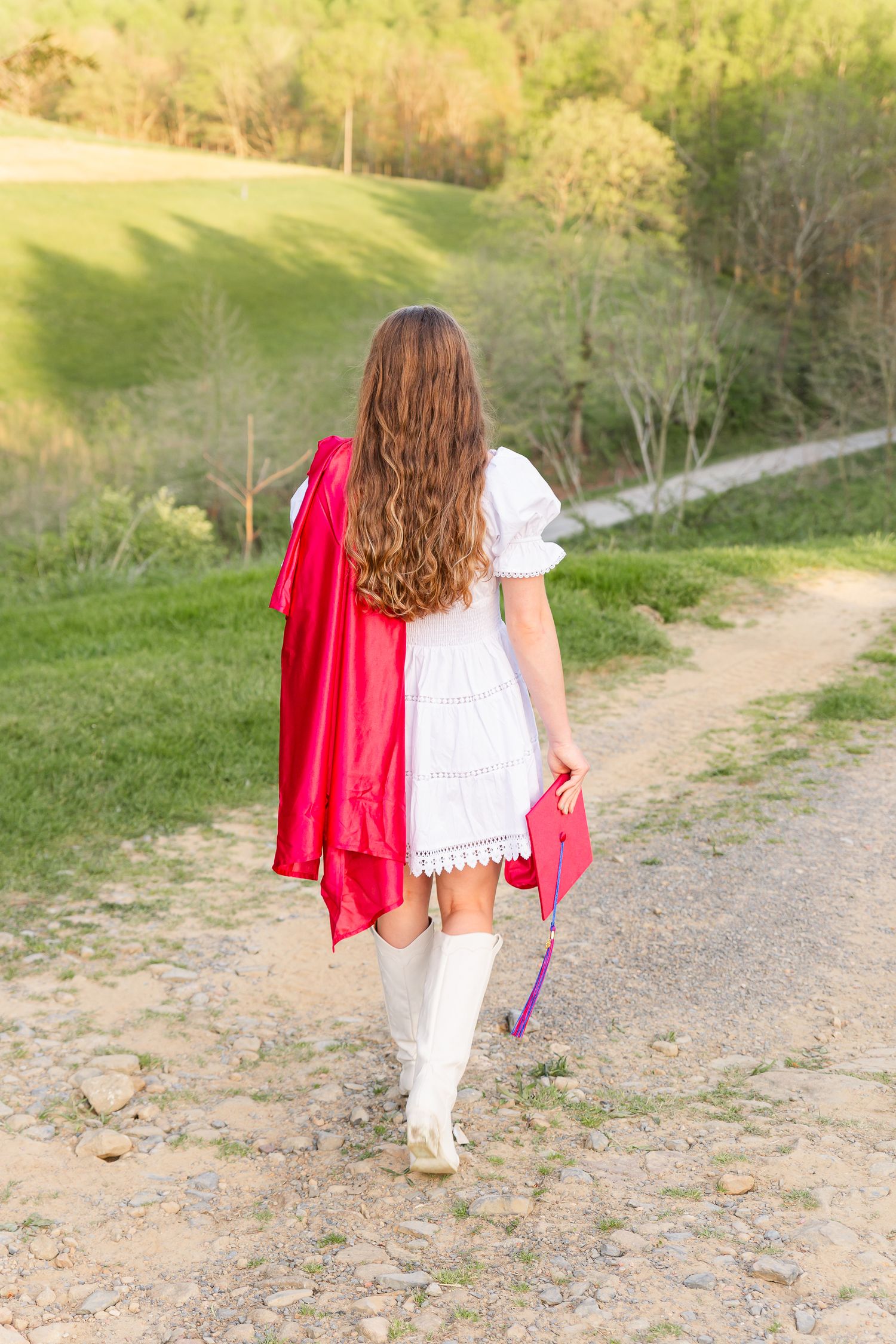 Graduate in white dress and red cap walks along path carrying graduation regalia.