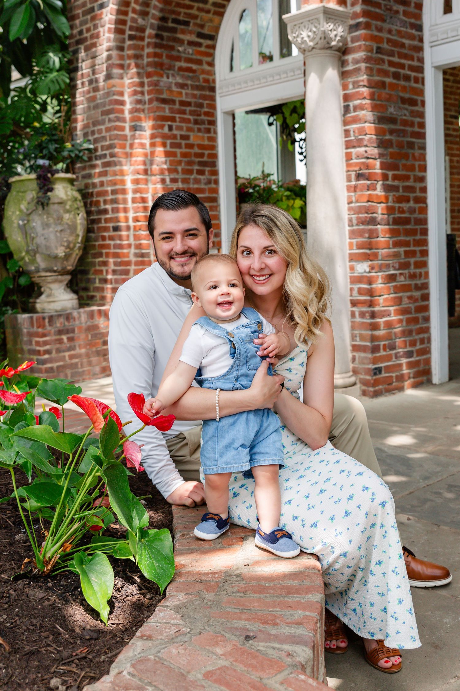 Young family with baby boy posing on brick steps surrounded by red flowers and lush greenery.