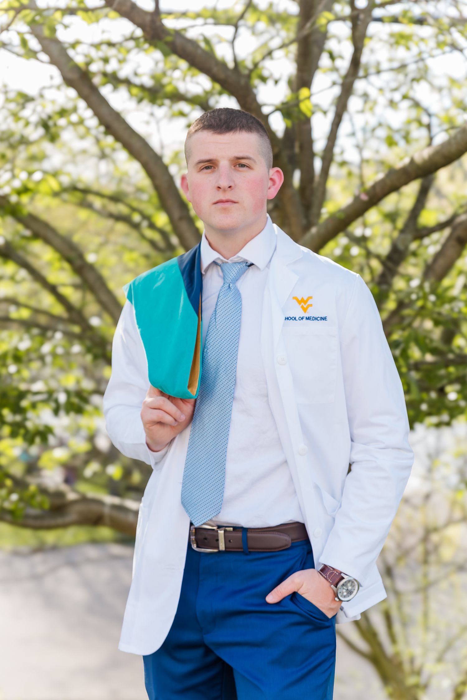 Student in white shirt and blue tie poses with graduation attire outdoors.