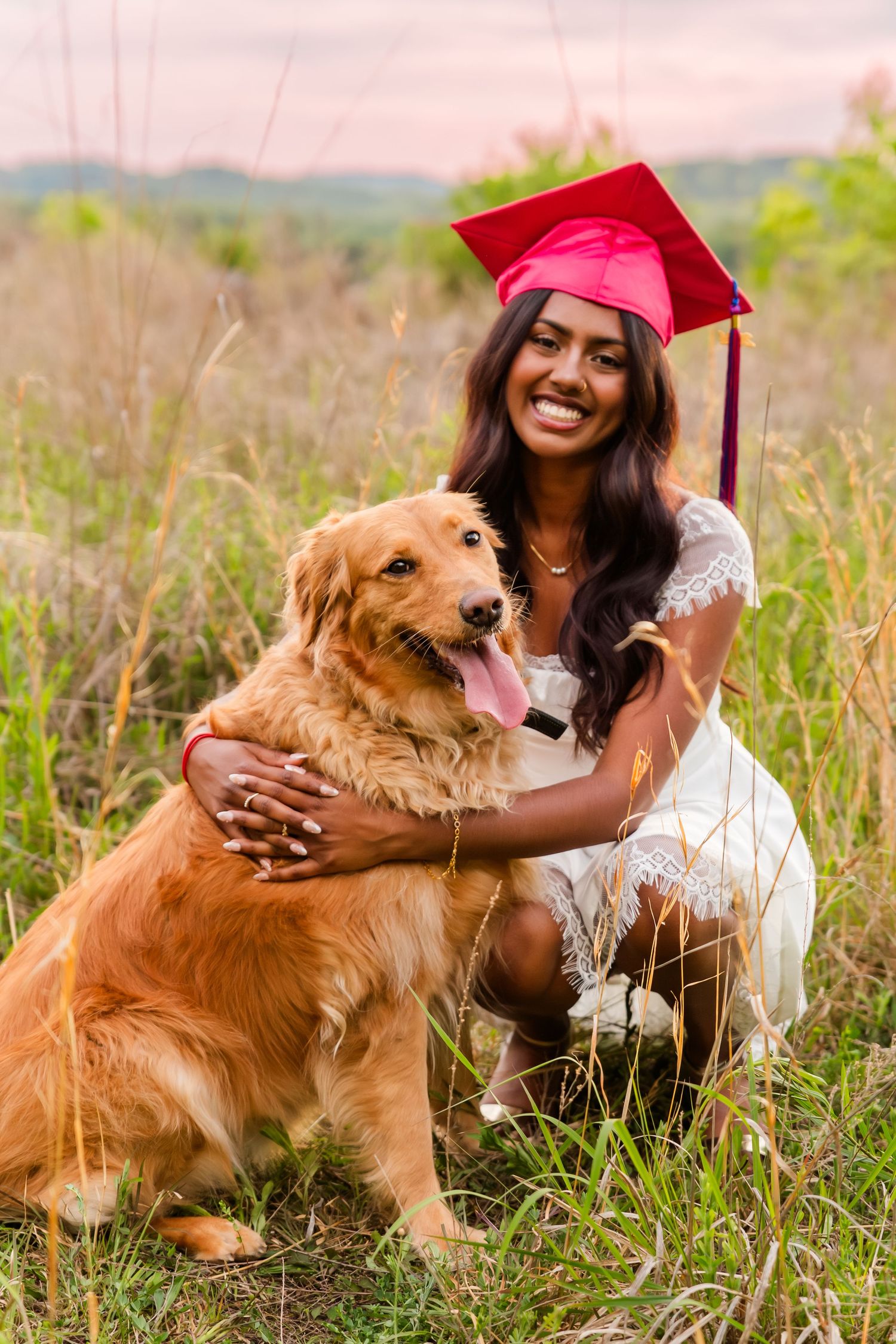 Graduate in red cap poses with golden retriever dog in sunny meadow setting.
