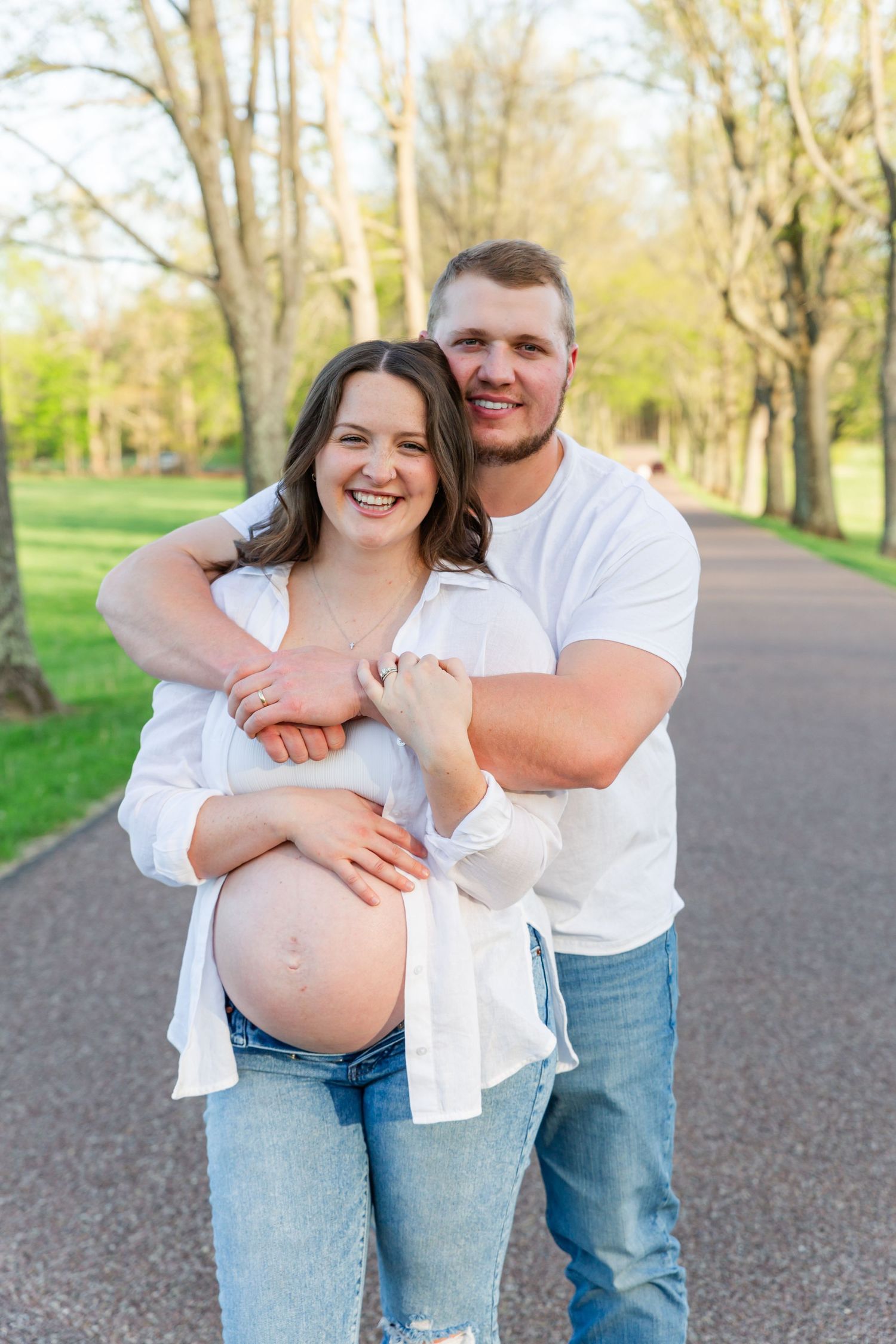 A maternity photo of a couple embracing on a tree-lined path in springtime.