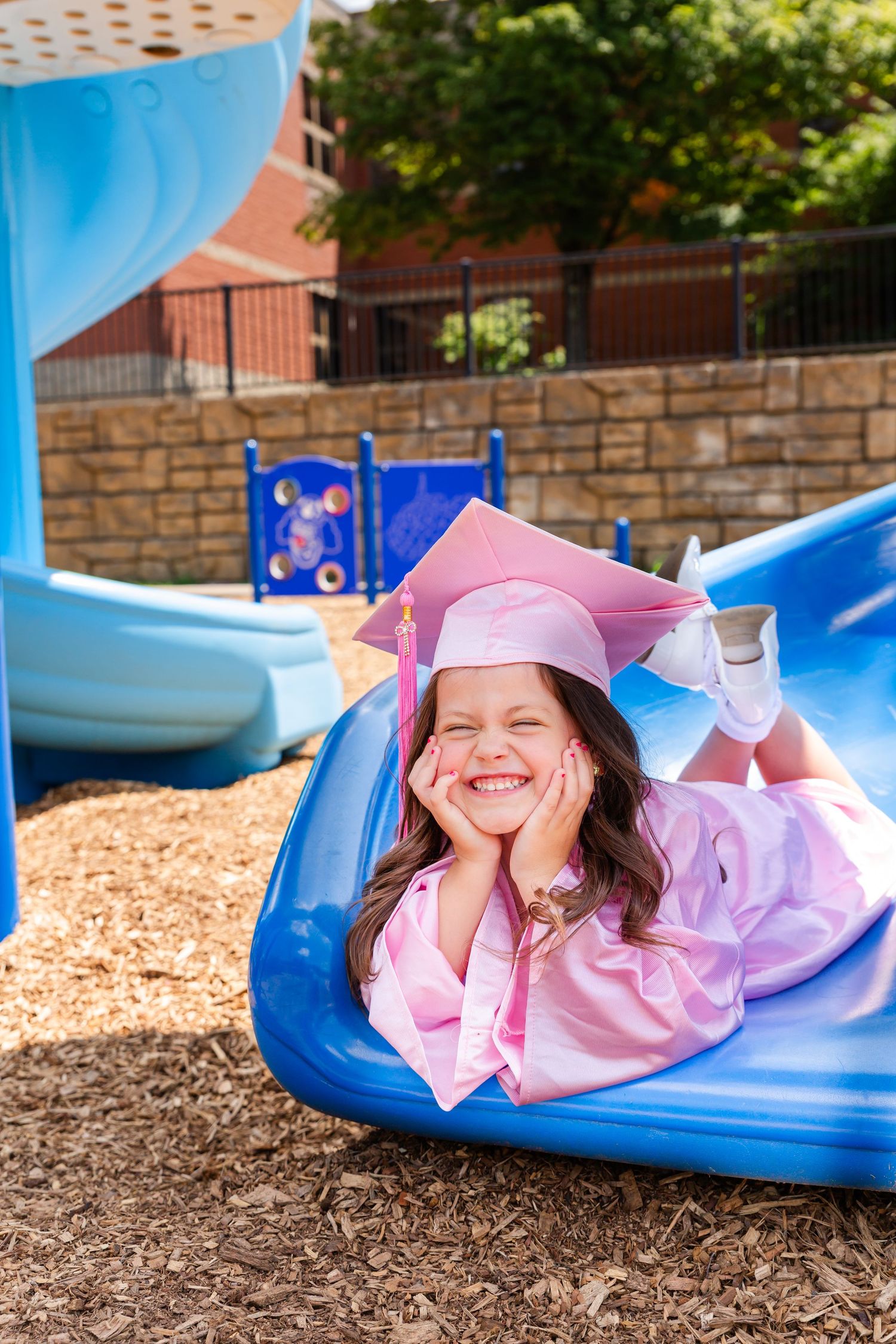 Playful graduation photo on blue playground slide with pink cap and gown.