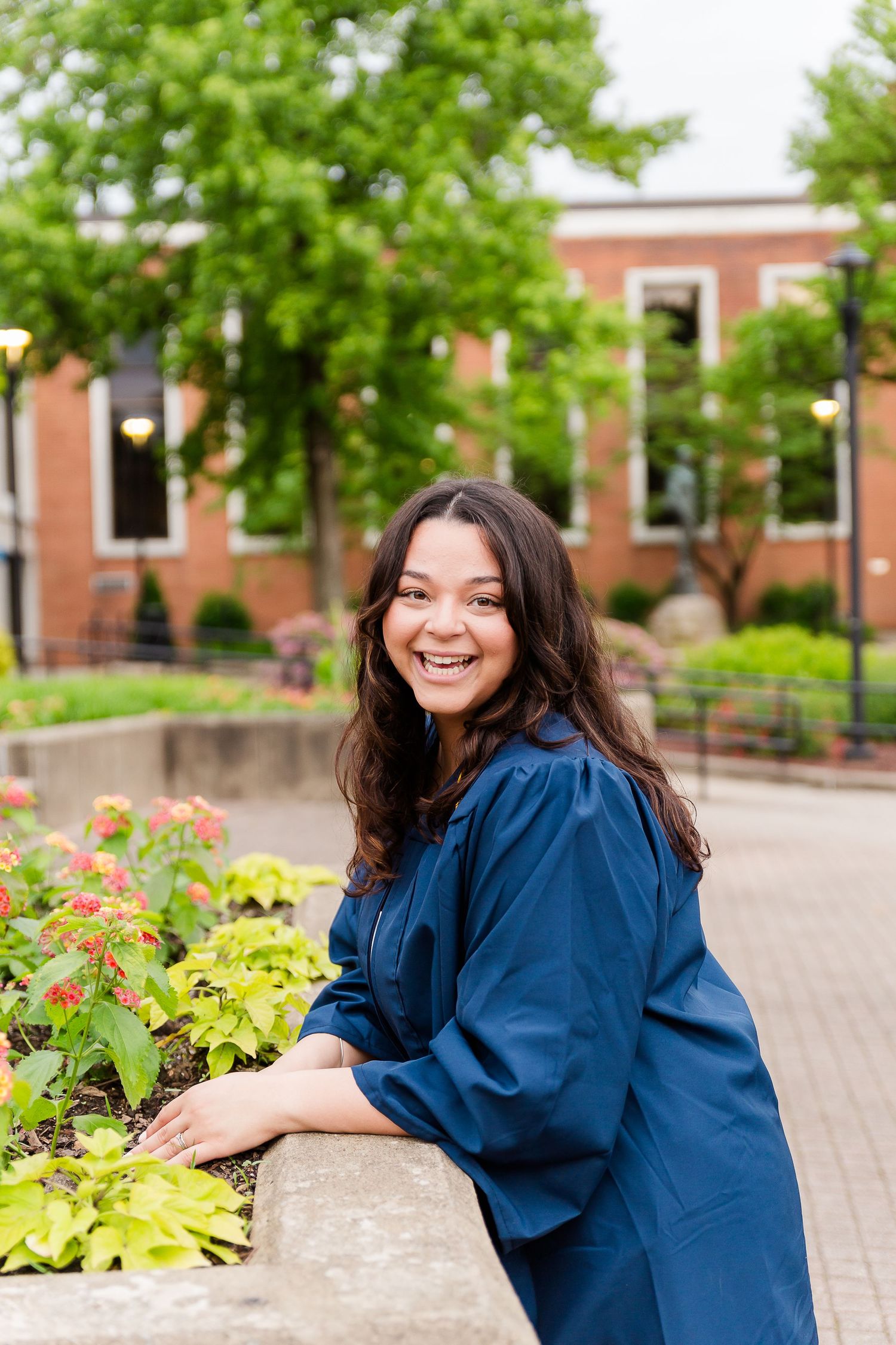 Graduate in blue cap and gown poses near colorful garden plants on campus walkway.