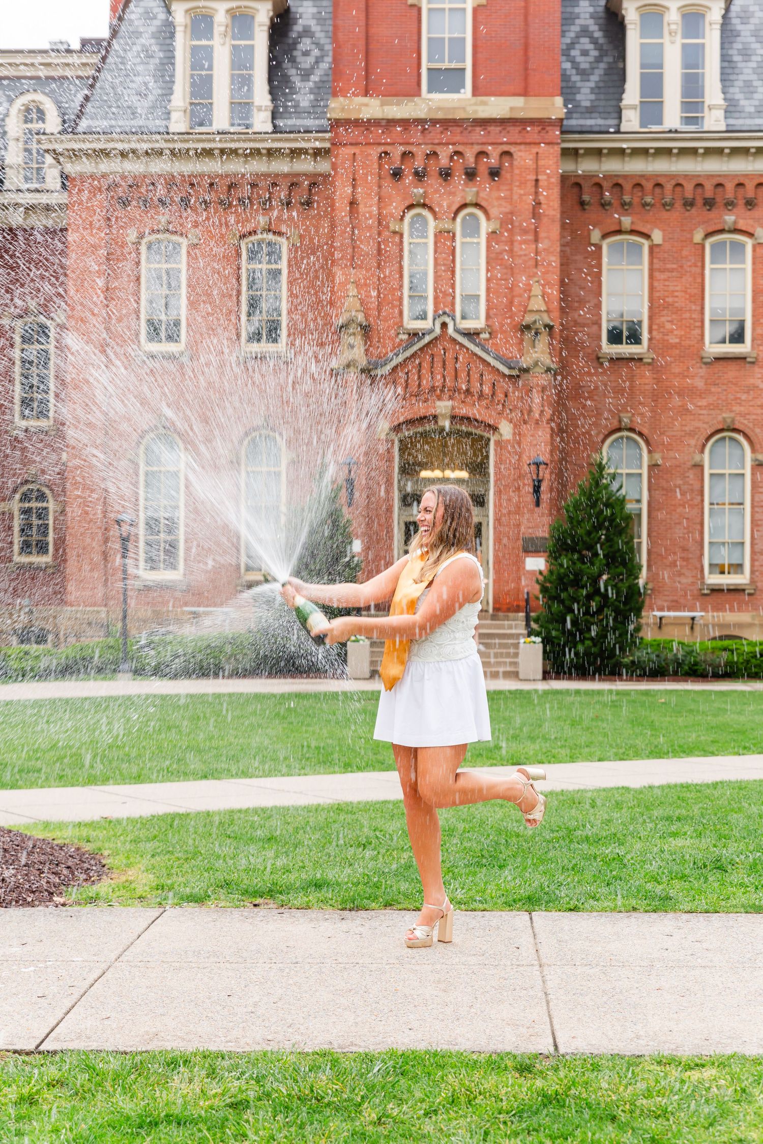 Graduate in white dress plays in sprinkler on sunny day in front of historic red brick university building.