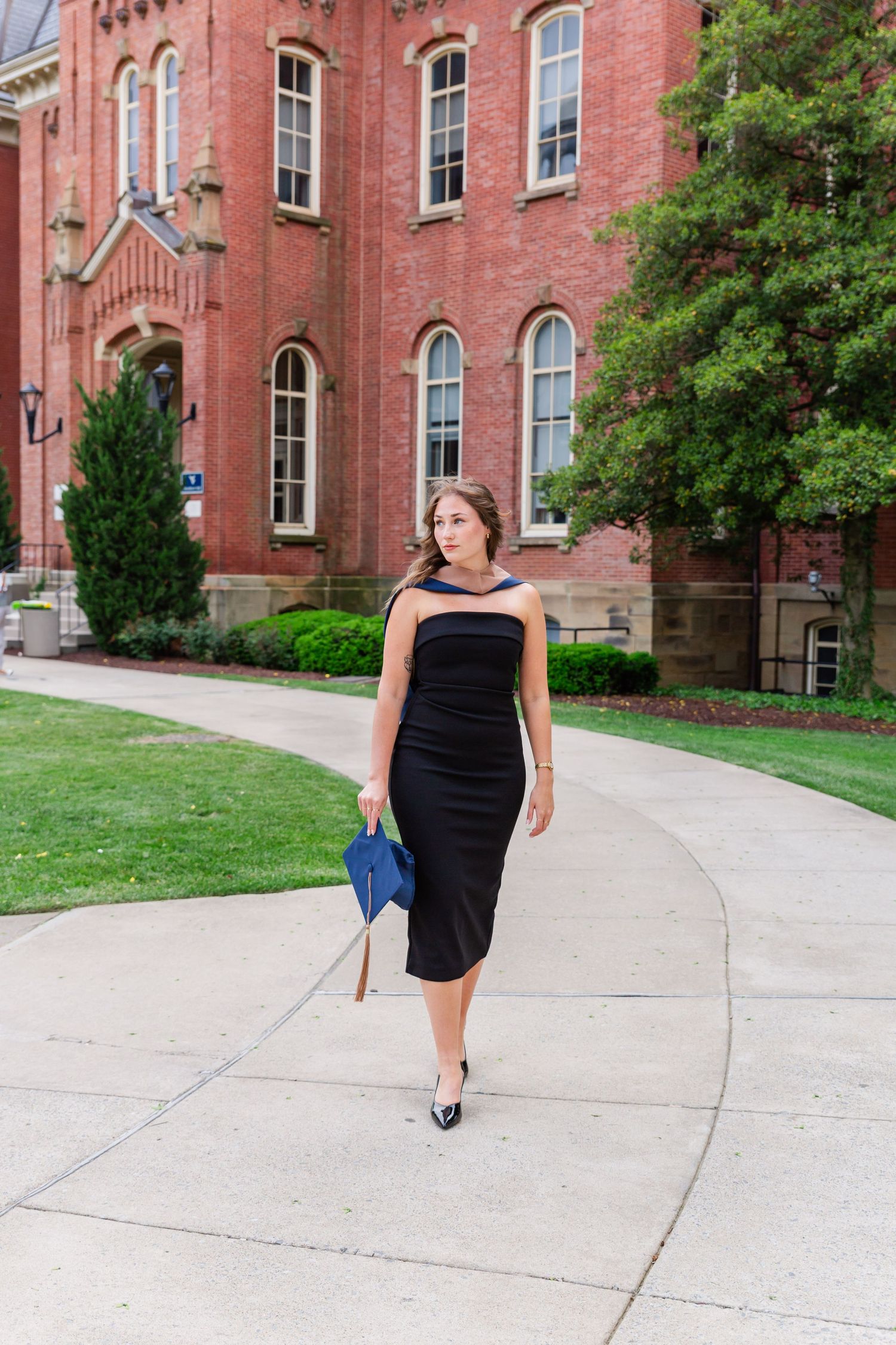 Graduate in black dress walks confidently along campus sidewalk near historic brick building.