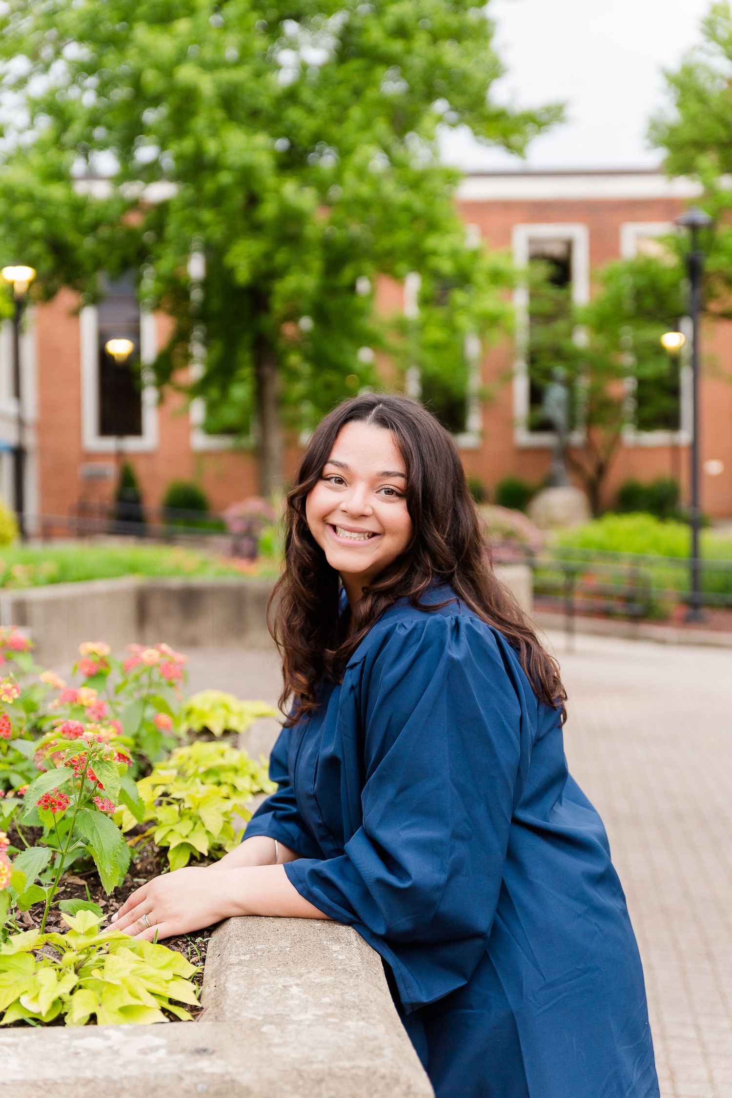 Graduate in blue gown poses near colorful garden beds on campus walkway.