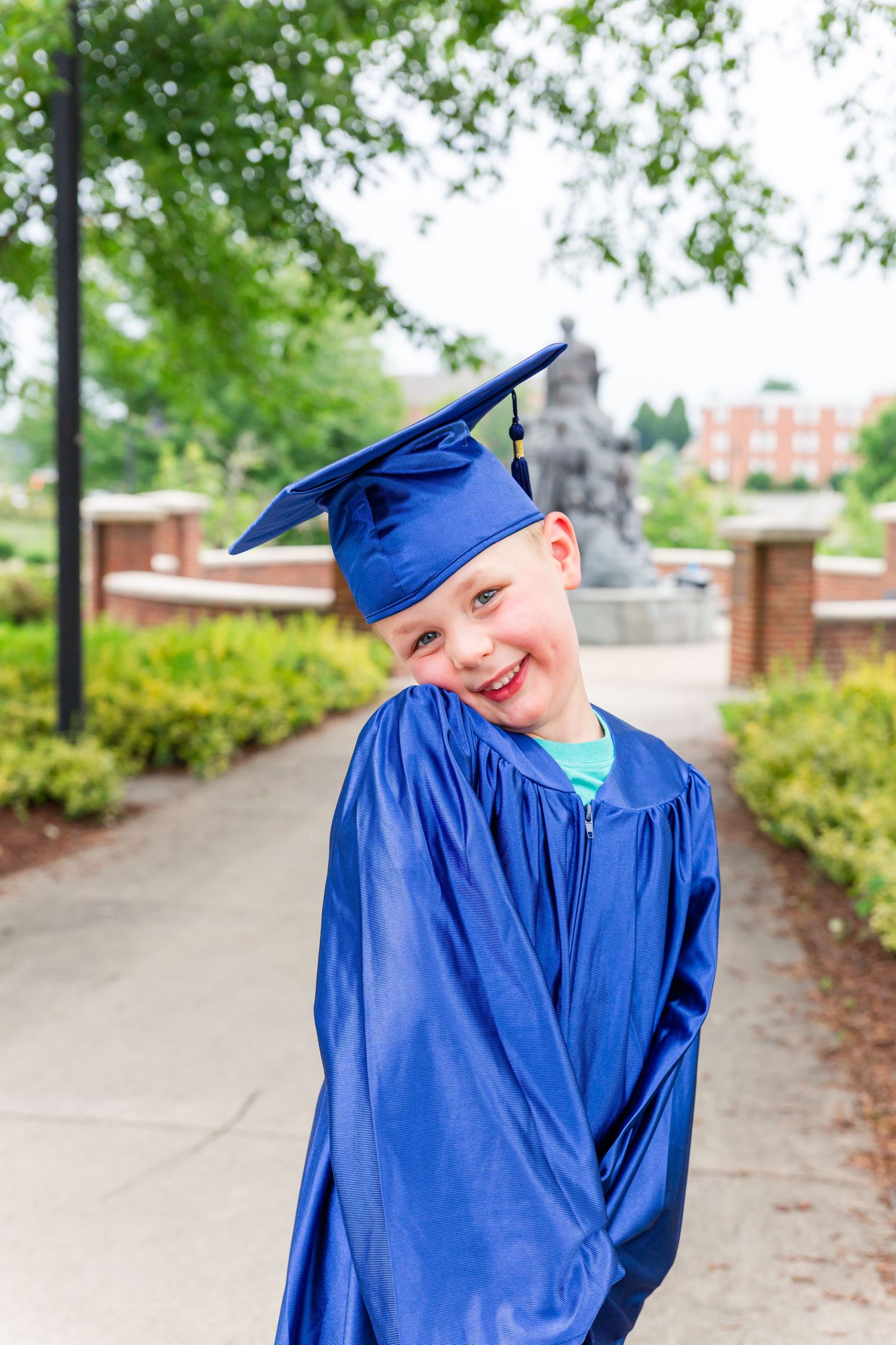 Graduate in blue cap and gown strikes playful poses on campus pathway.