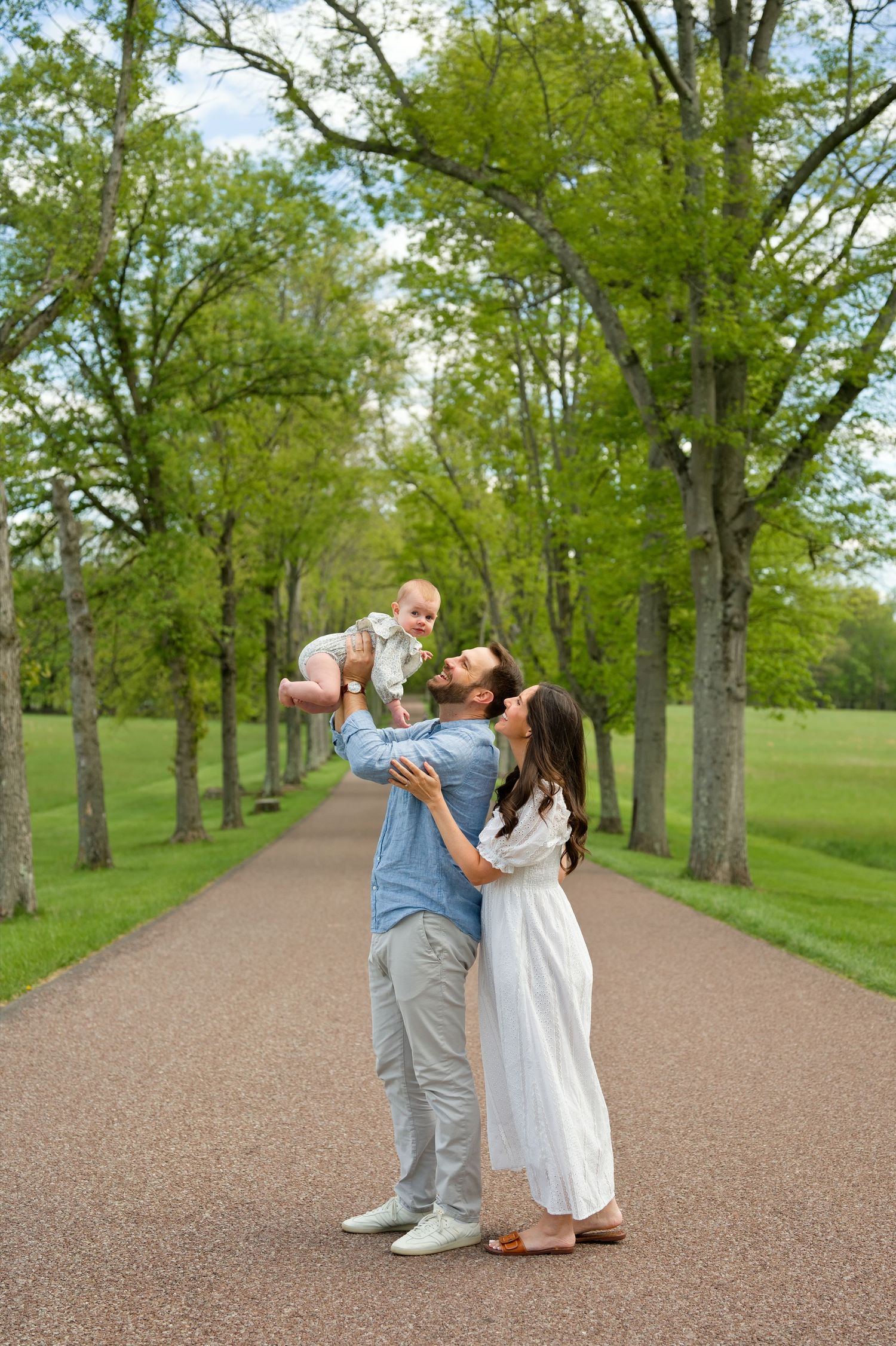 A happy family poses on a tree-lined path, father lifting baby while mother in white dress looks on.