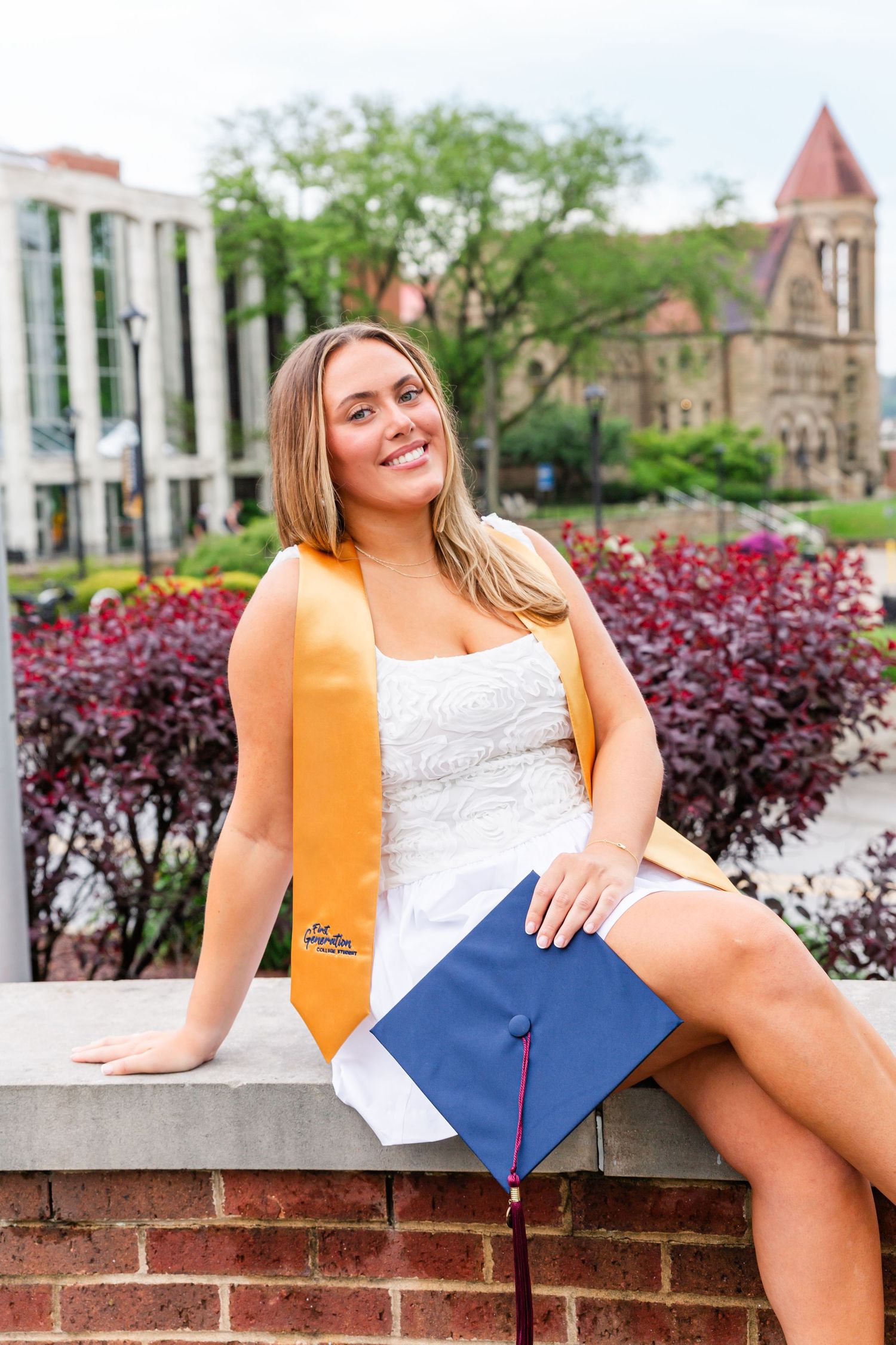 Graduate in white dress and gold stole poses with diploma on brick wall near campus flowers.