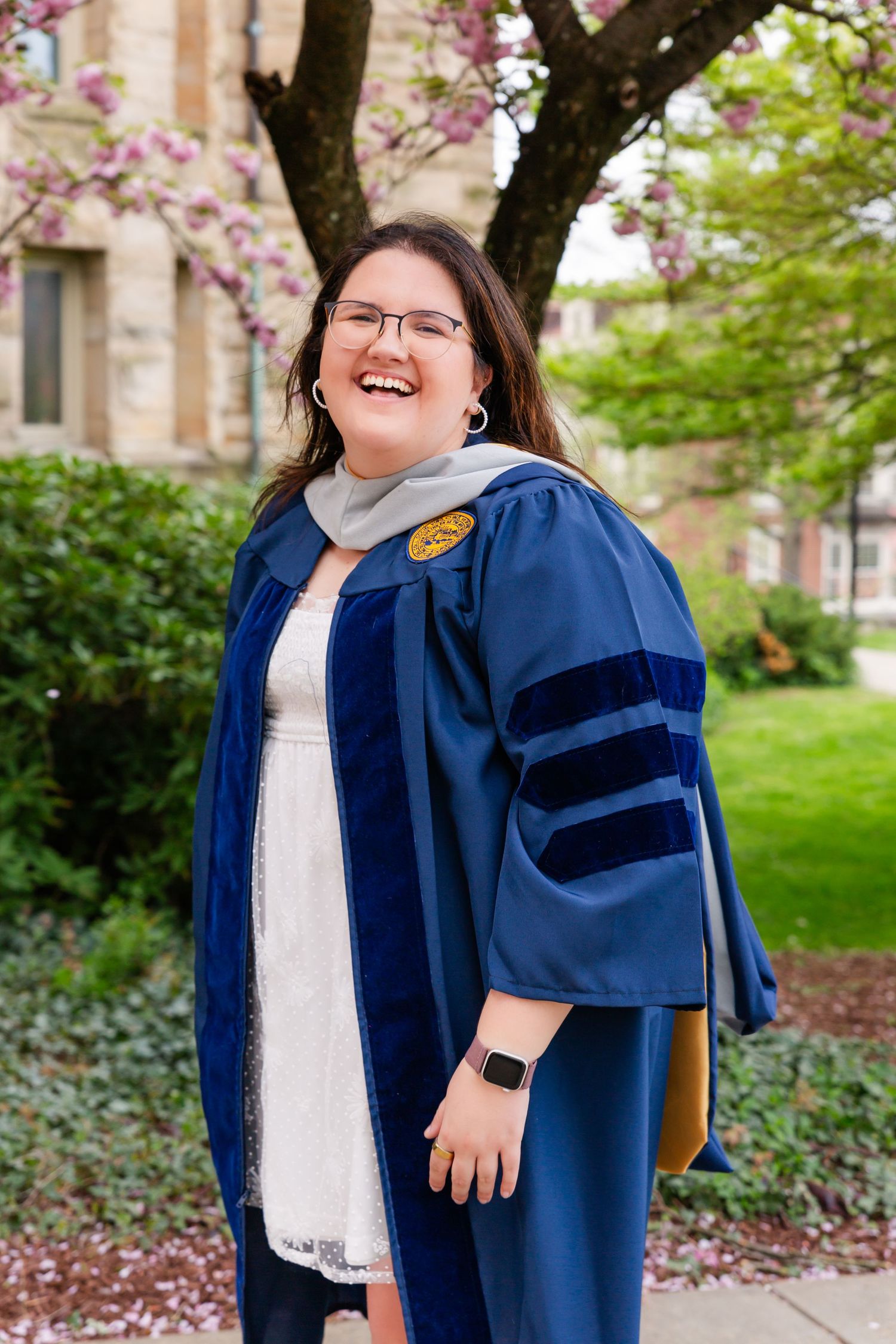 Graduate in blue academic regalia poses outdoors on campus with brick buildings and trees in background.