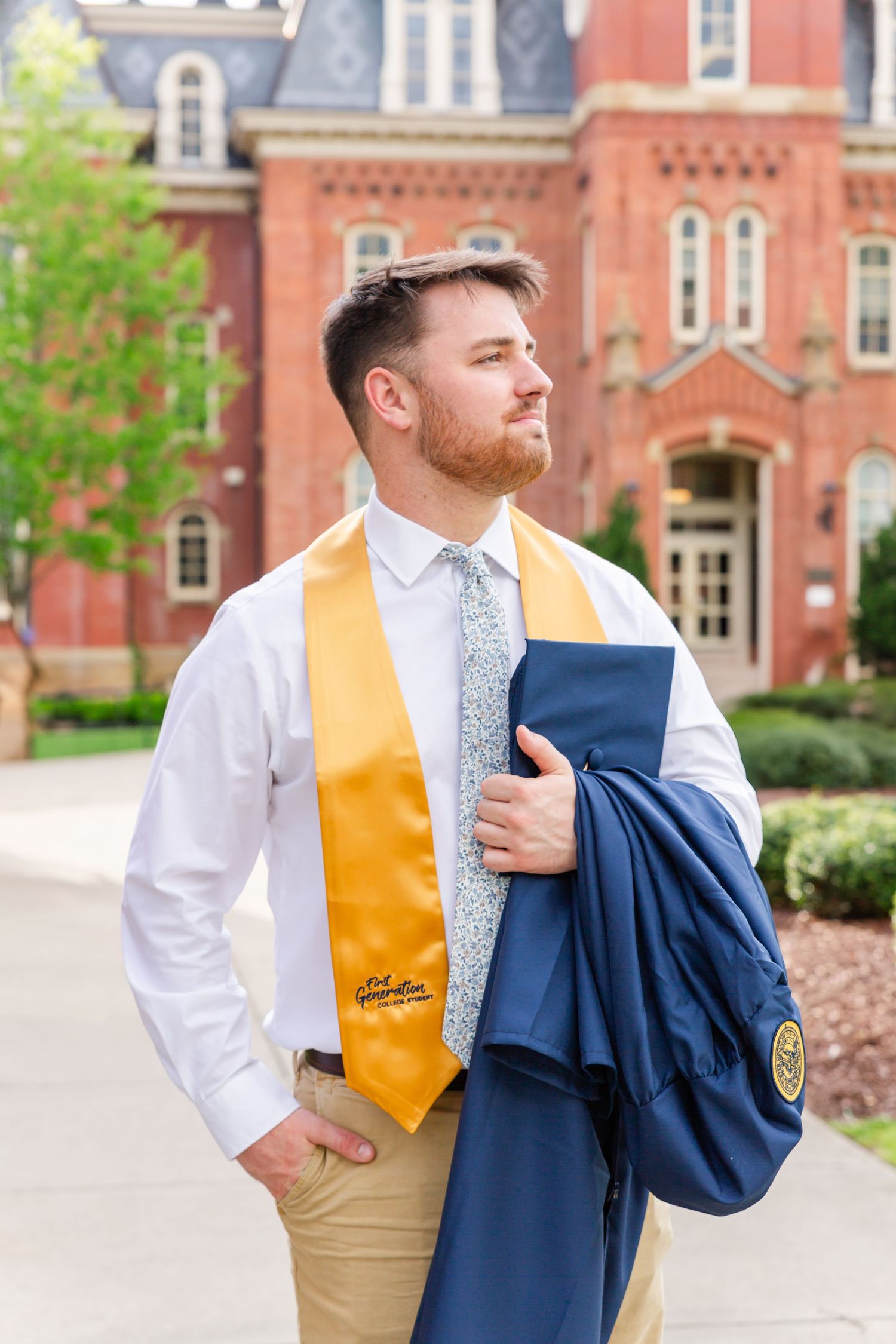 Graduate in formal attire with gold stole poses in front of historic university architecture.