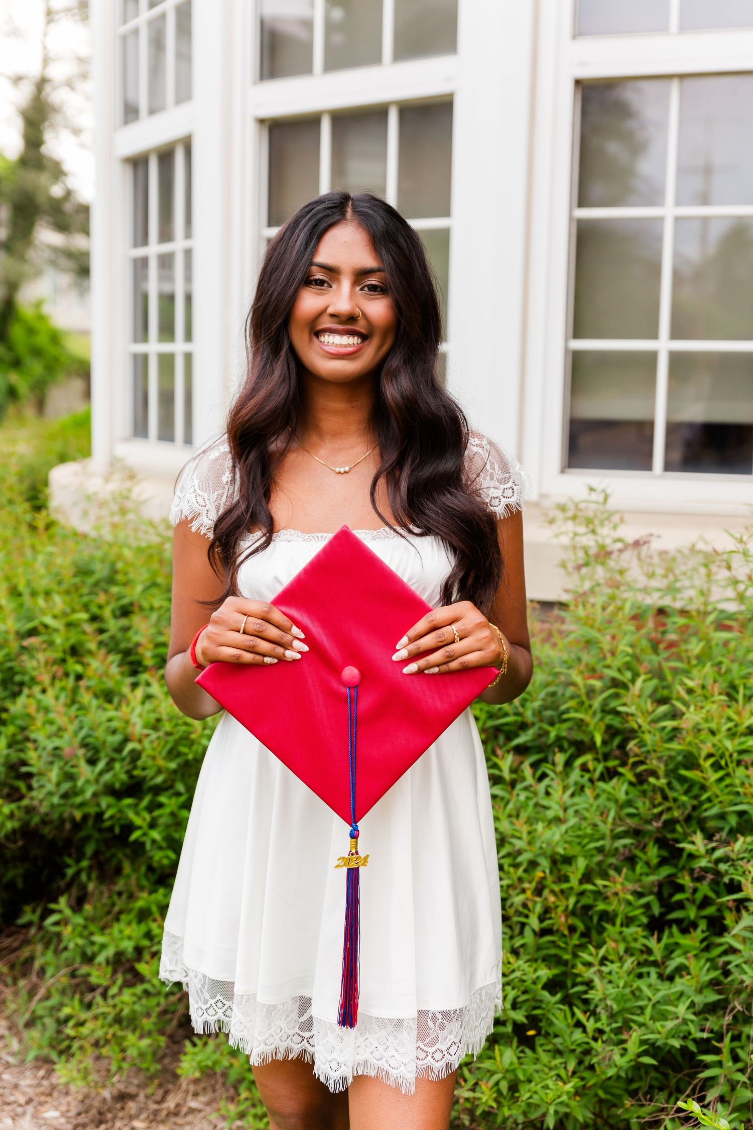 Graduate in white lace dress holds red graduation cap while standing in front of window with greenery.