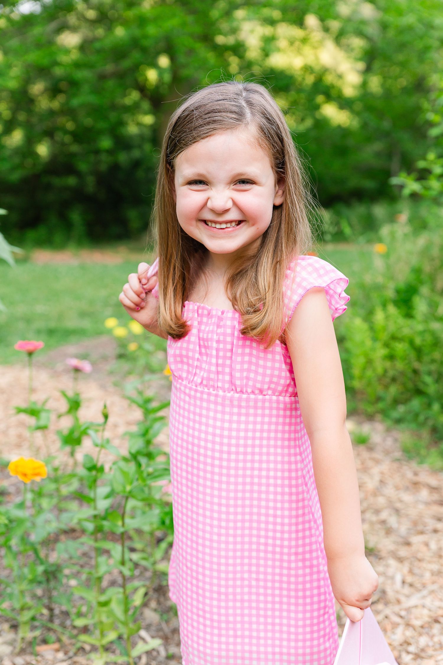 Child in pink gingham dress poses playfully in a garden setting.