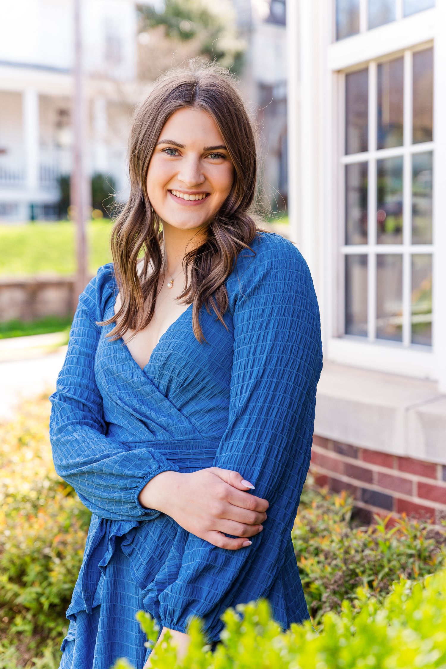 Smiling young woman in a blue wrap dress posing outdoors near a white building and green shrubs.