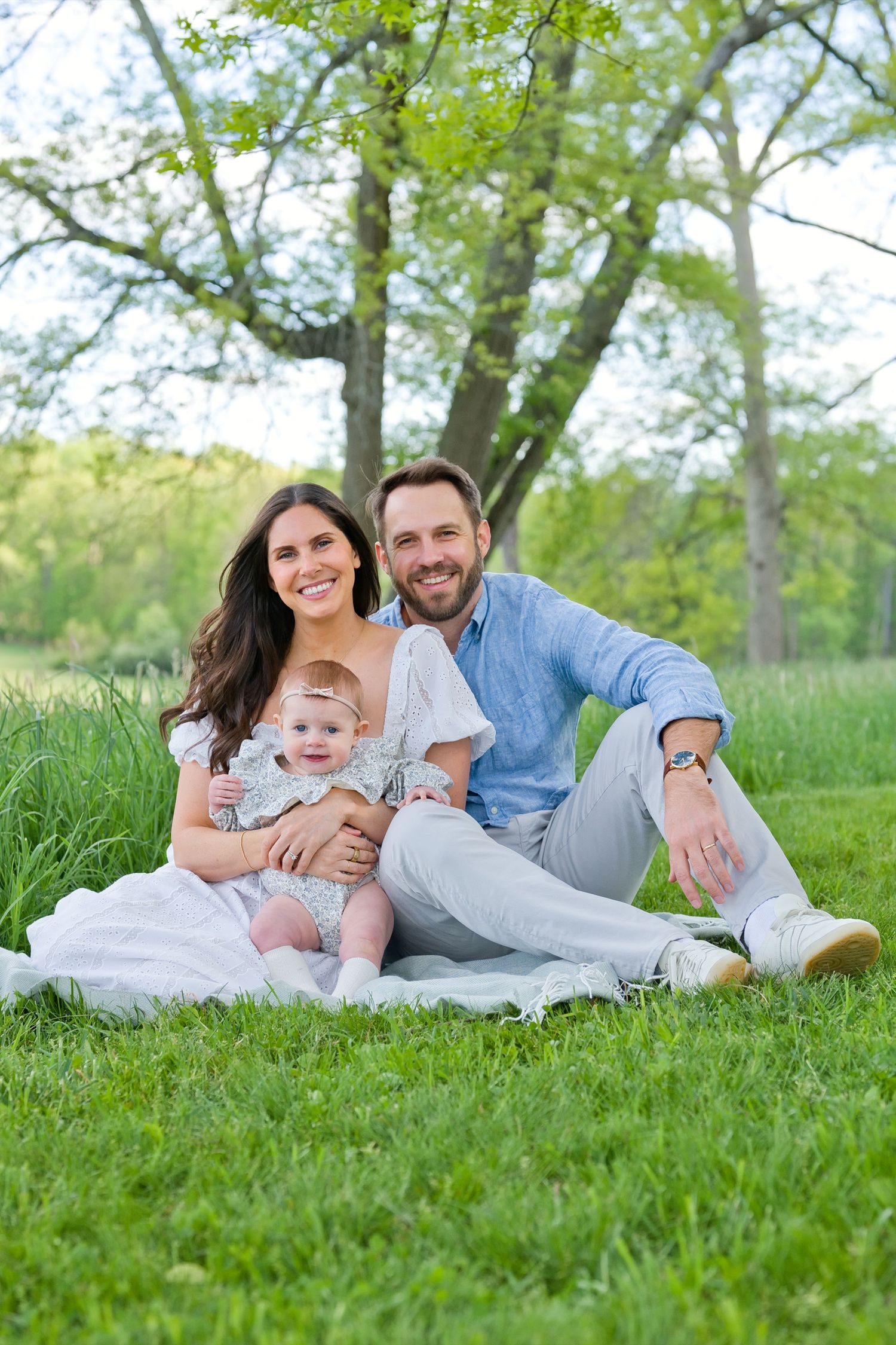 A happy young family with a baby sits on a blanket in a lush green park under a large tree.