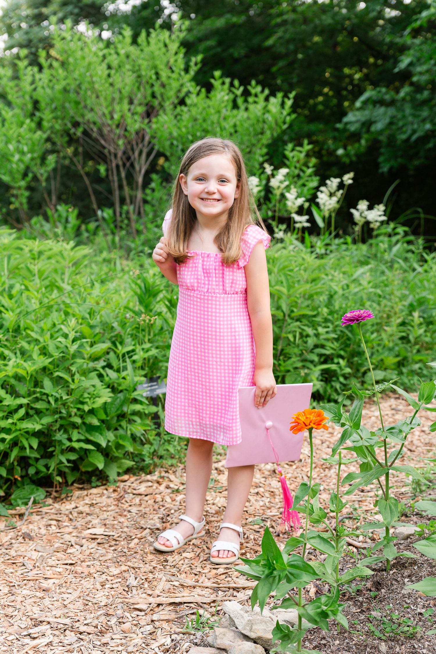 A pink gingham dress stands out against lush garden greenery with bright orange zinnias blooming in a mulched flower bed.