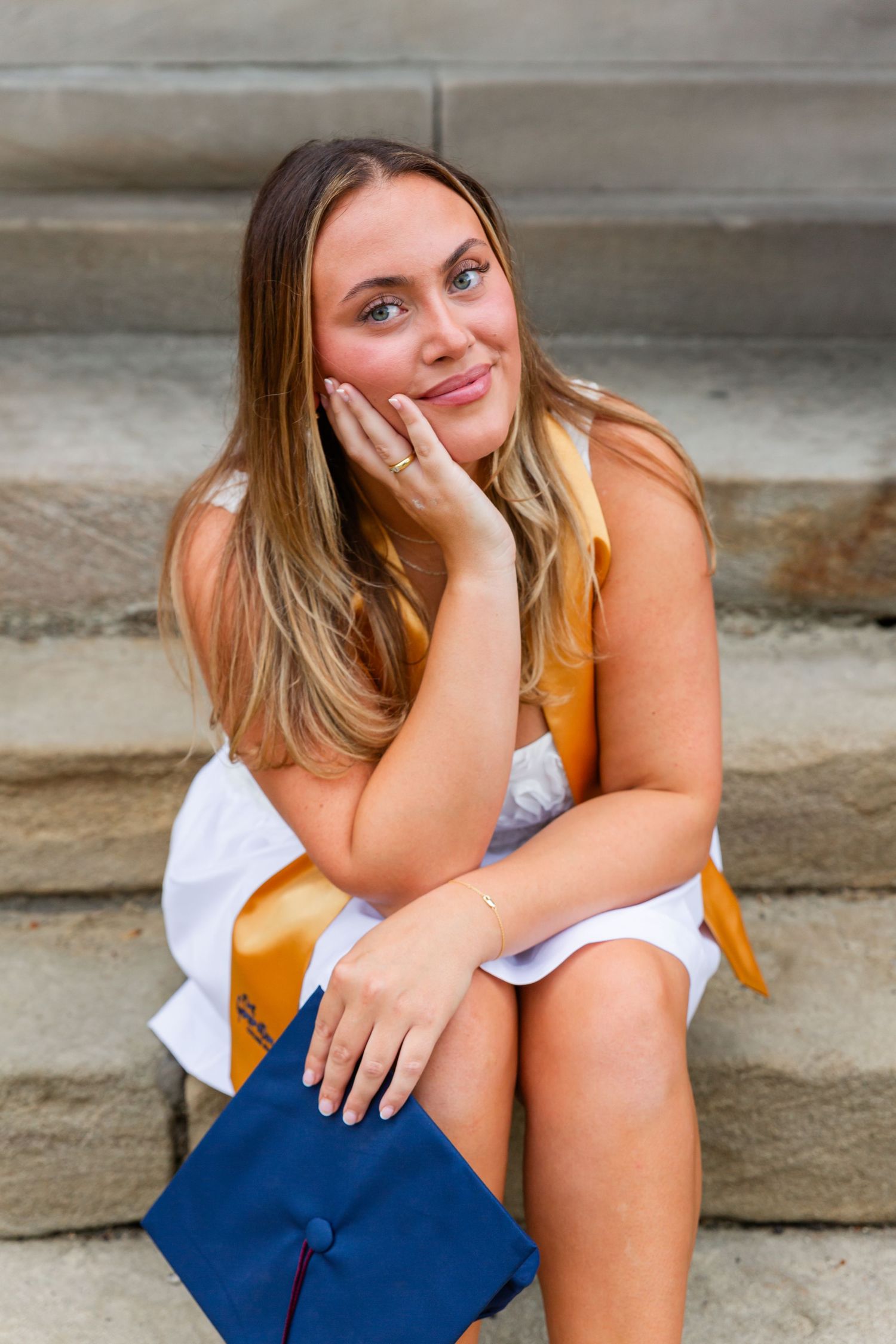 Graduate in white dress and gold stole sitting casually on stone steps.
