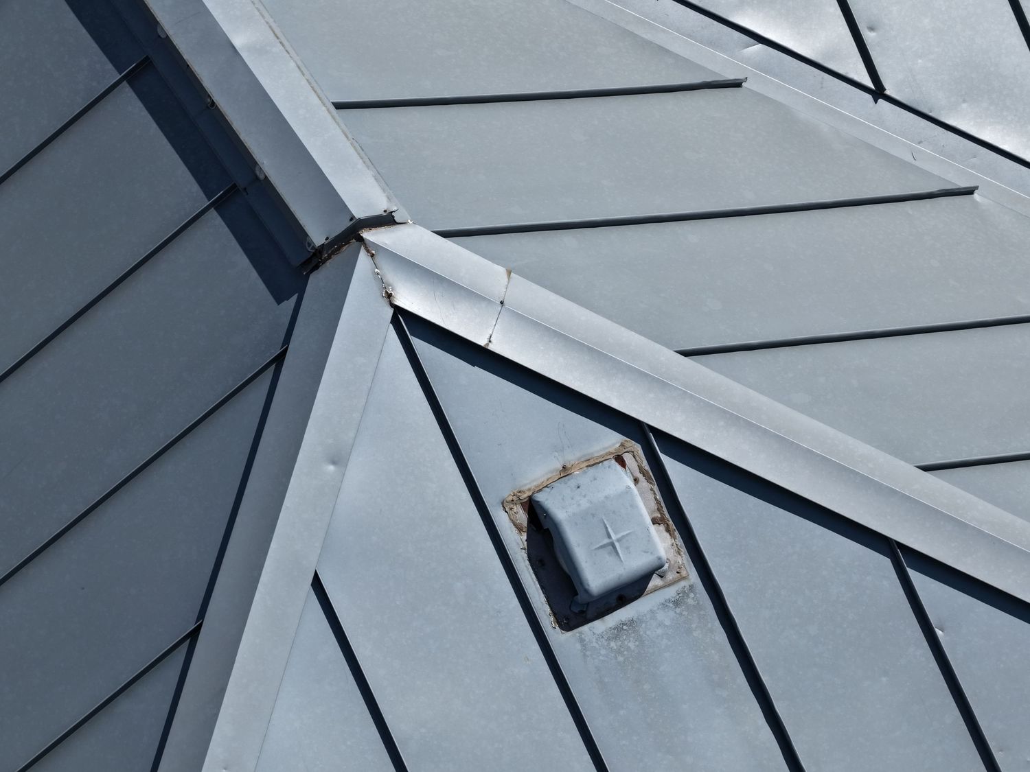 Aerial close-up of a gray metal roof and a small roof vent at the intersection of slopes.