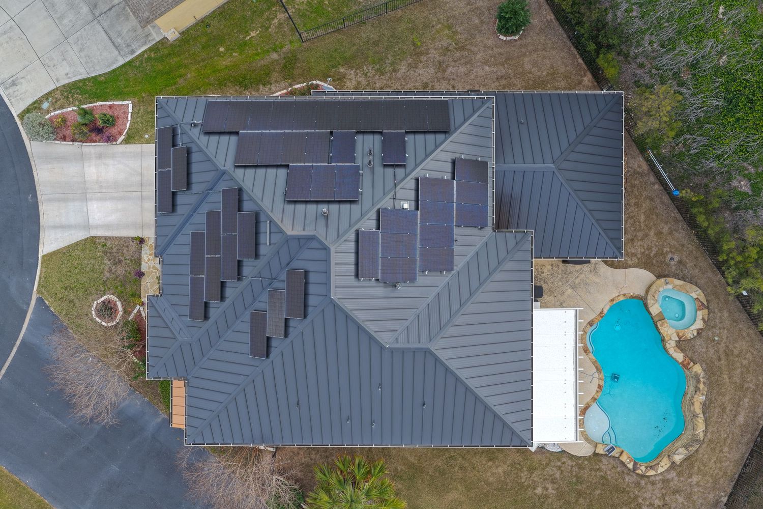 Aerial view of a home with solar panels on a gray metal roof, featuring a kidney-shaped pool in the backyard.