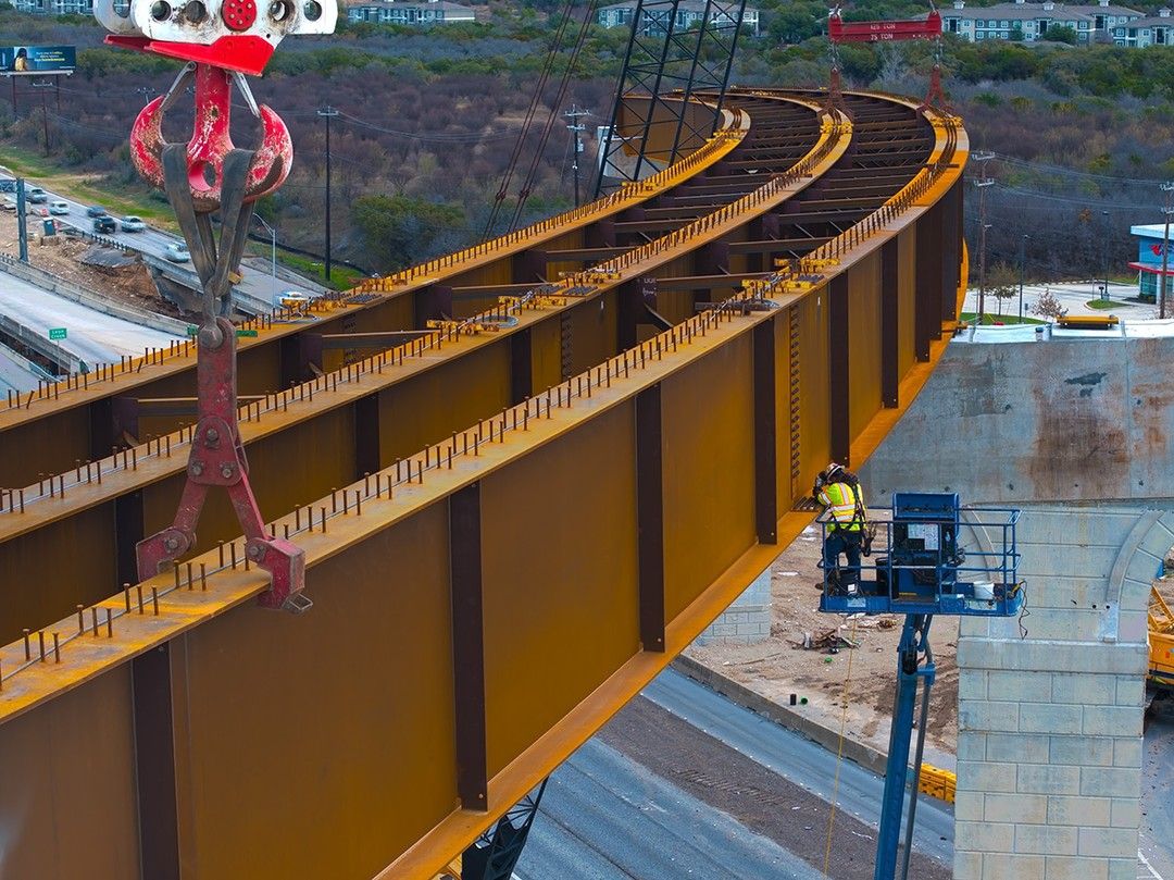 Aerial view of curved fly-over bridge construction with orange steel girders and worker on lift platform.