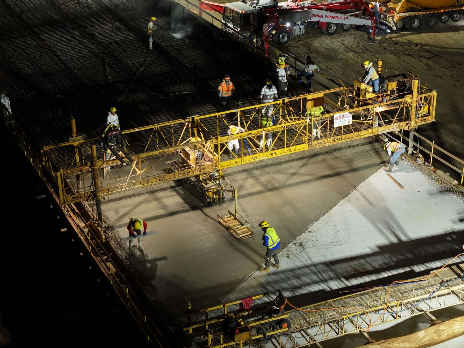 Construction workers pouring and leveling concrete at night using a yellow Bid-Well on a bridge site.