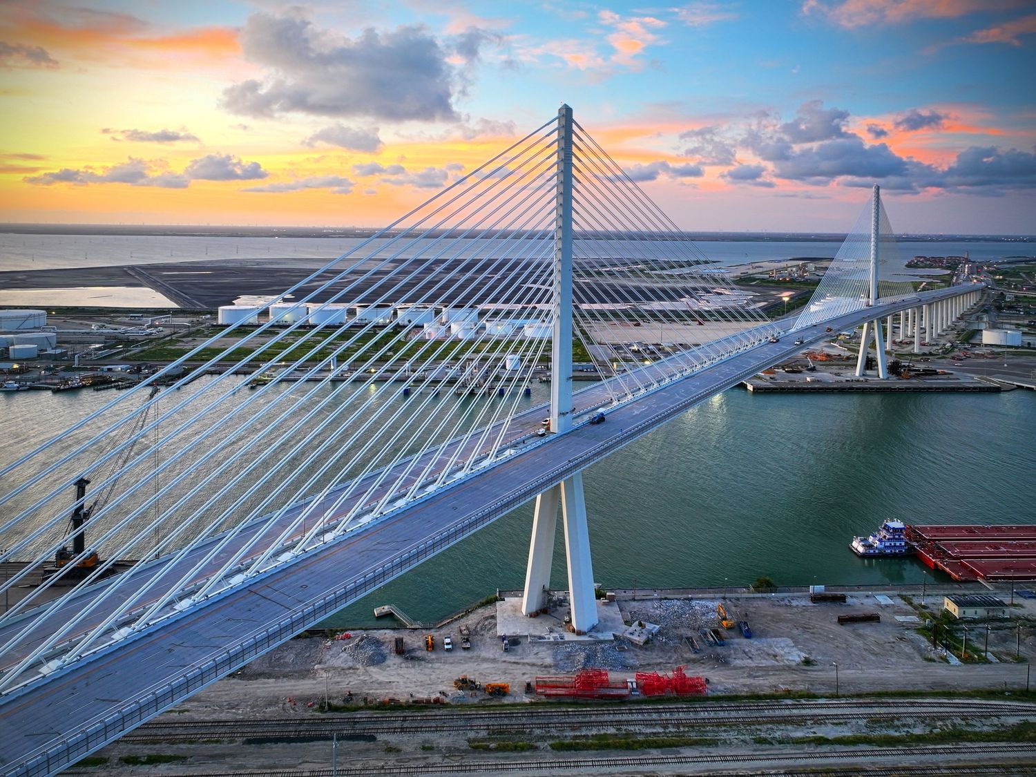 Aerial view of a modern cable-stayed Corpus Christi's harbor bridge at sunset over calm waters near an industrial port.
