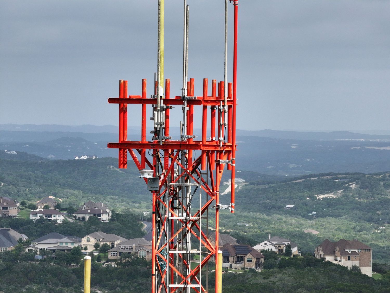 Red cell tower with antennas stands tall above a scenic green hillside suburban landscape under overcast skies.