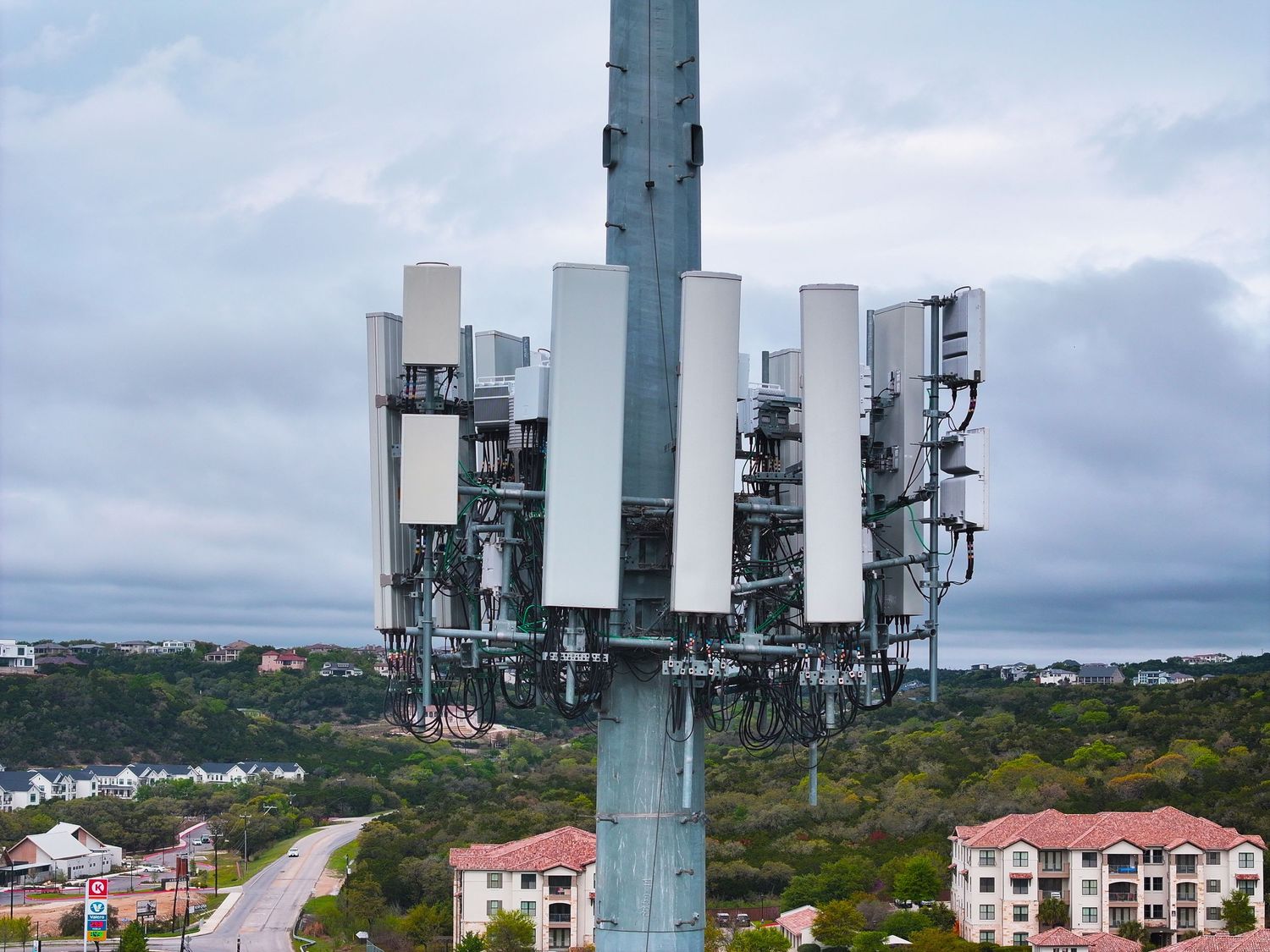 Aerial view of a tall cell tower with multiple 5G antennas against a cloudy sky in a suburban area.