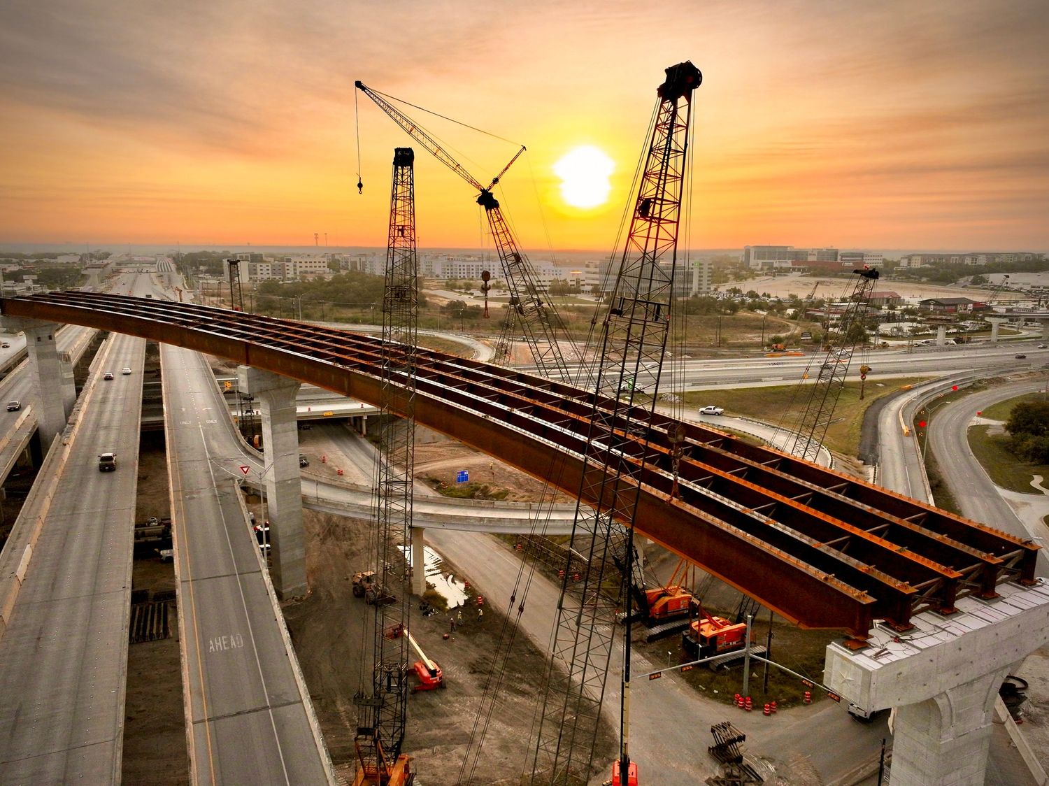 Aerial view of highway bridge construction at sunset with large cranes lifting steel girders over a busy interchange.