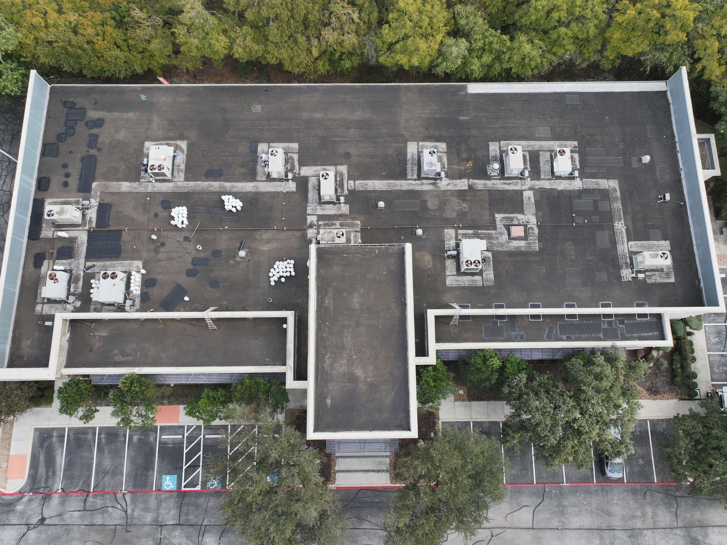Aerial drone view of a large commercial building rooftop with HVAC units, surrounded by trees and a parking lot.