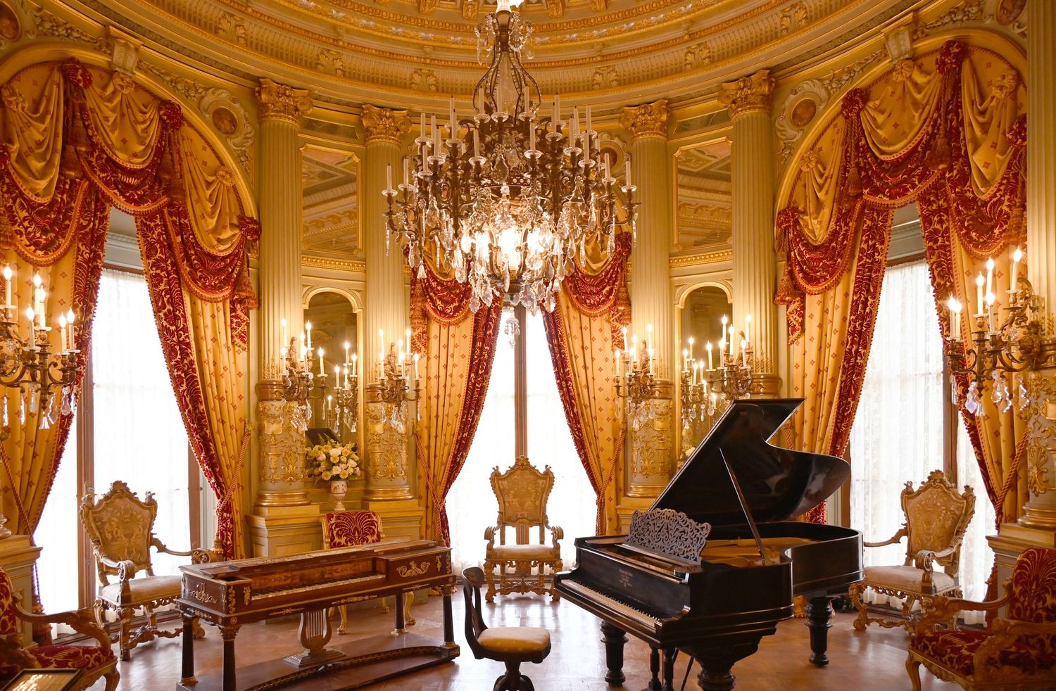 Gold Vanderbilt ballroom with grand piano, crystal chandelier, red velvet drapes, and ornate candelabras in a luxurious palace interior.