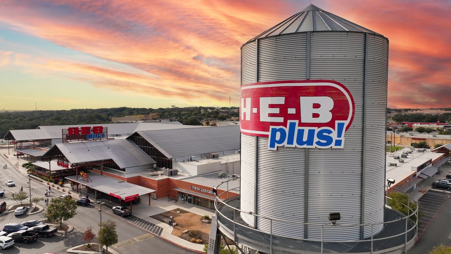 H-E-B Plus! store exterior with water tank signage at sunset, showcasing vibrant red and orange sky.