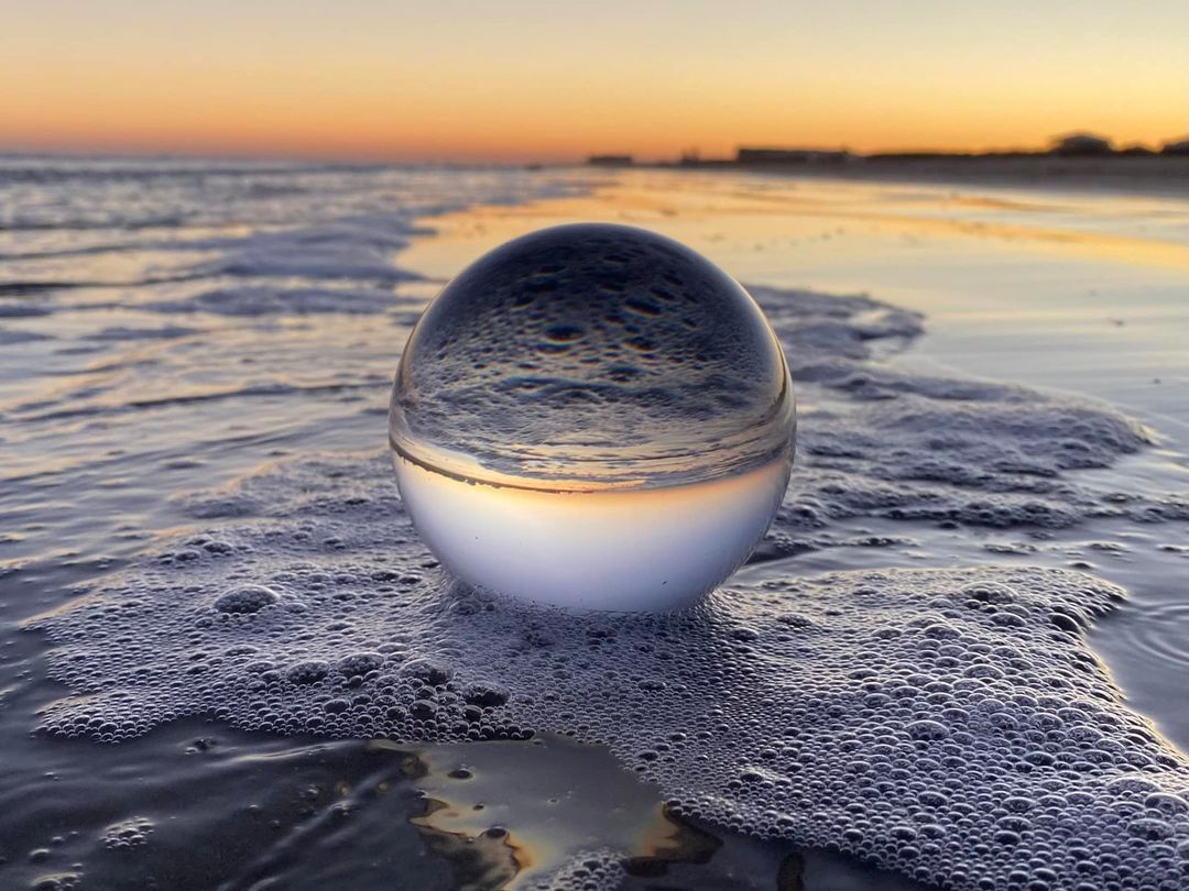 Crystal ball on sandy beach reflecting sunset sky and ocean waves in golden hour light.