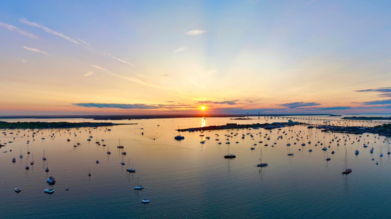 Aerial sunset view over a calm Newport harbor filled with sailboats, reflecting golden light on tranquil waters.