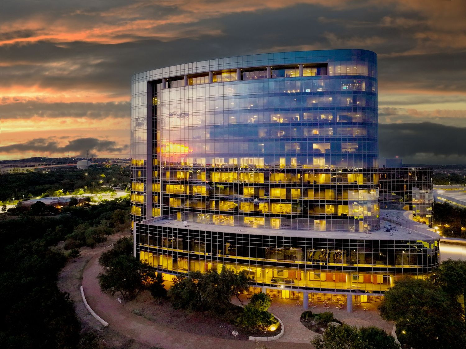 Modern glass office building illuminated at dusk with warm golden lights against a dramatic sunset sky.