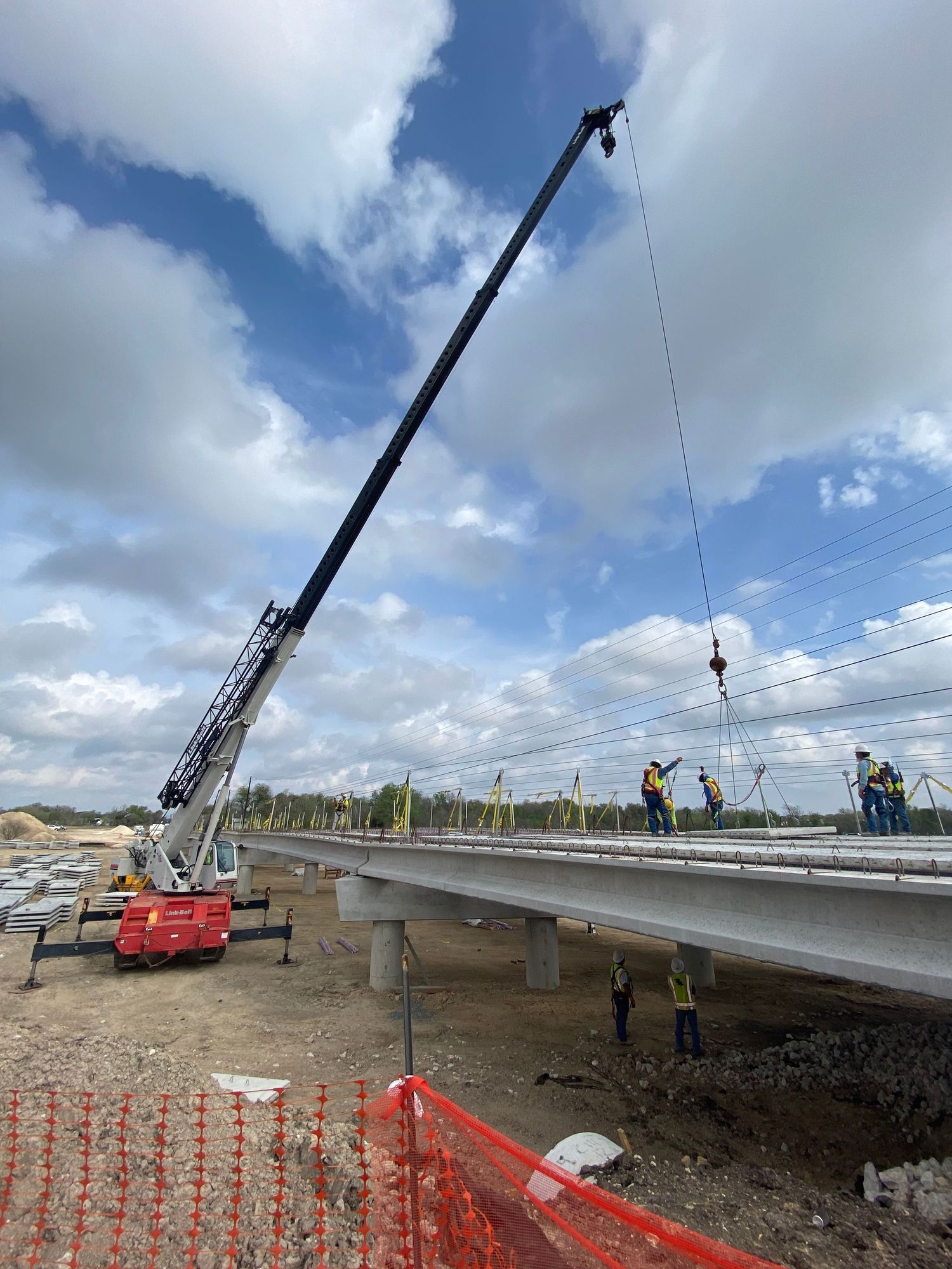 Construction crane lifting deck panels onto a roadway bridge with workers in safety vests on a cloudy day.