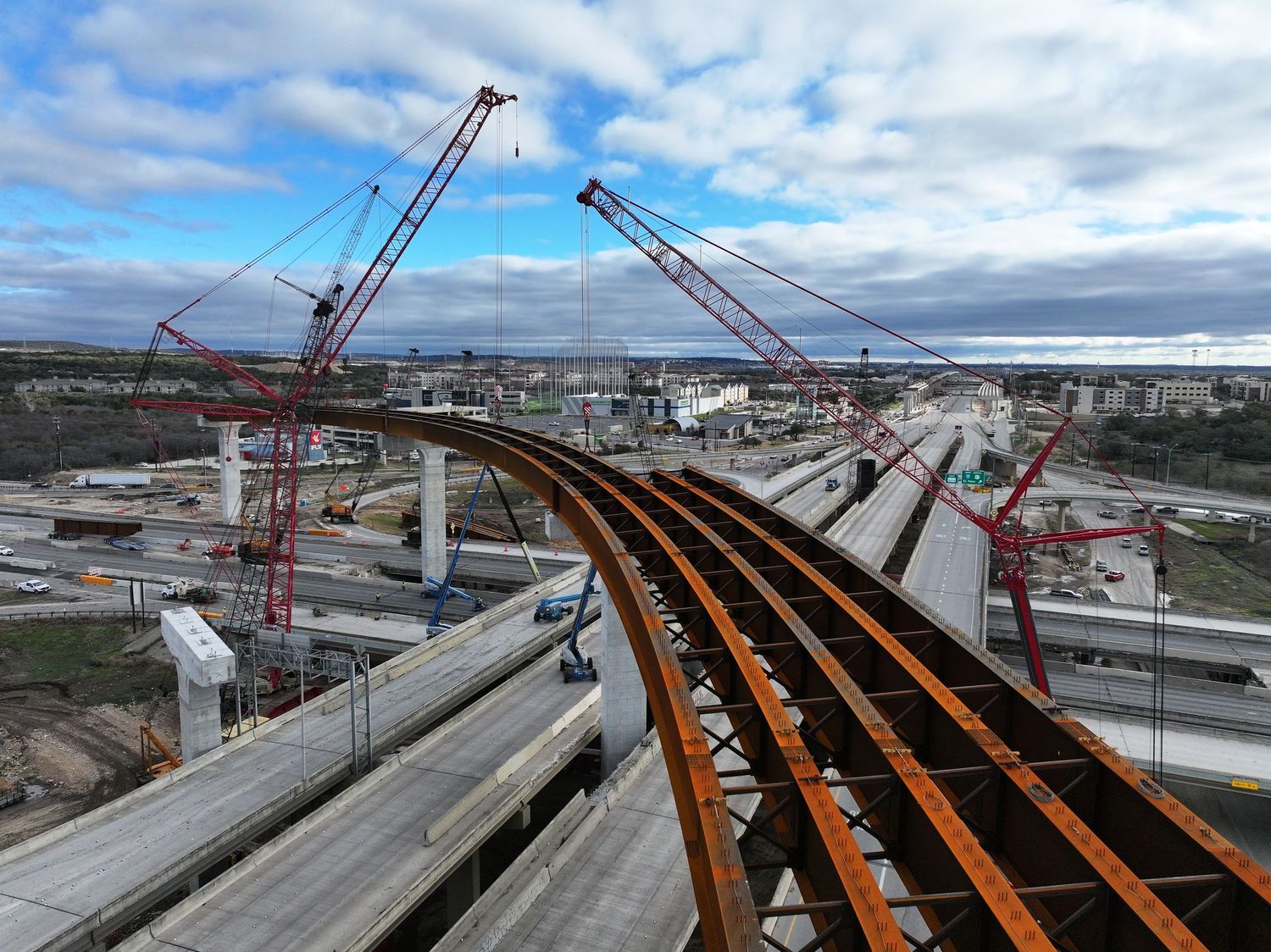 Aerial view of curved steel girders fly-over construction with red cranes against cloudy sky at the I-10 & 1604 interchange.