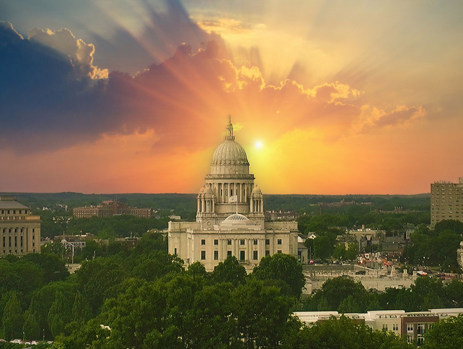 Rhode Island State Capitol building at sunset with dramatic golden rays and clouds over lush green trees.