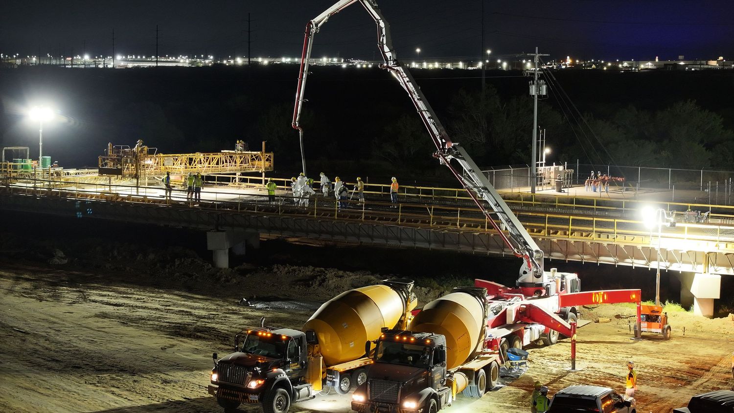 Night construction site with concrete pump truck and cement mixer pouring bridge deck under bright floodlights.