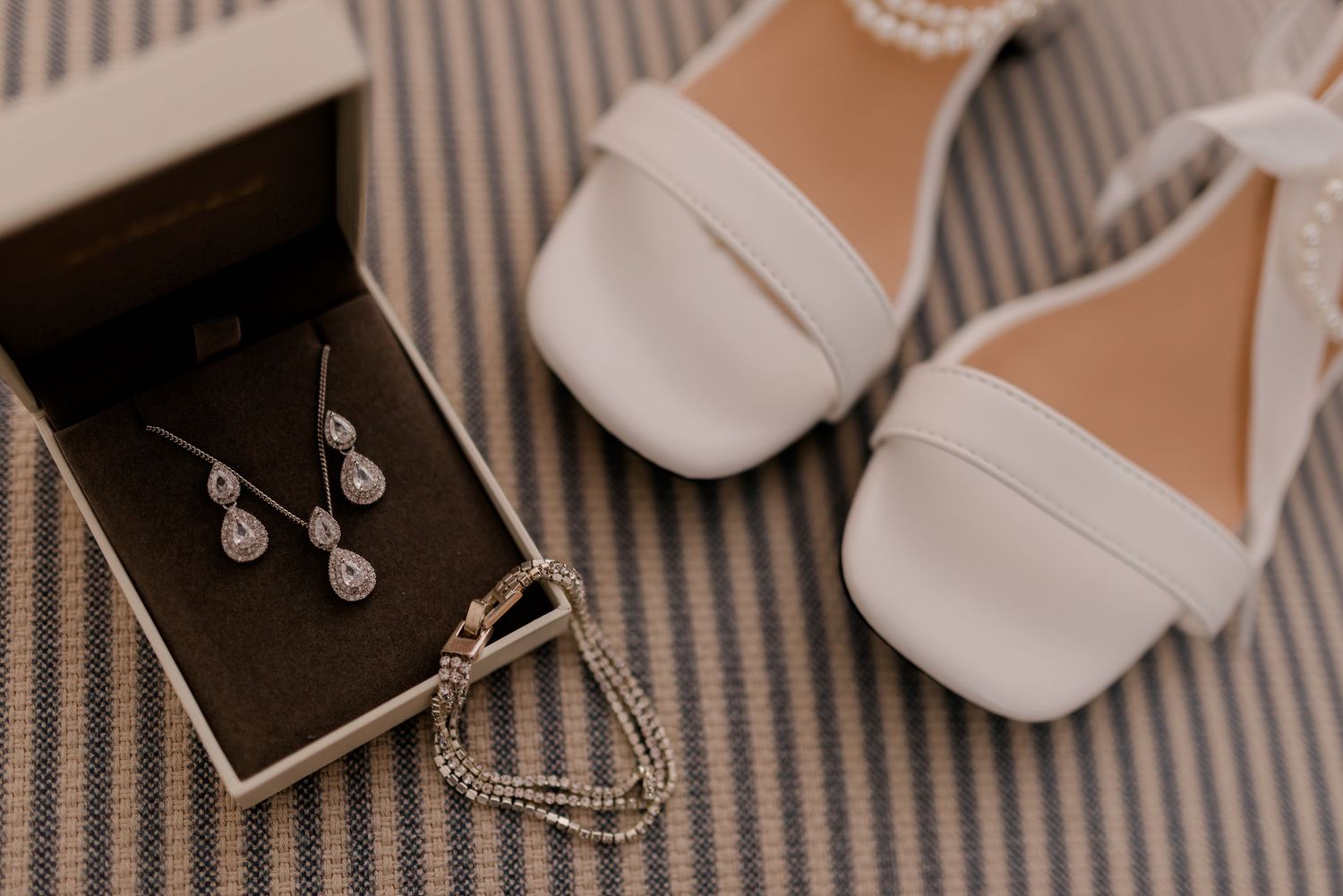 Elegant bridal accessories including white sandals and jewelry arranged on a striped surface.
