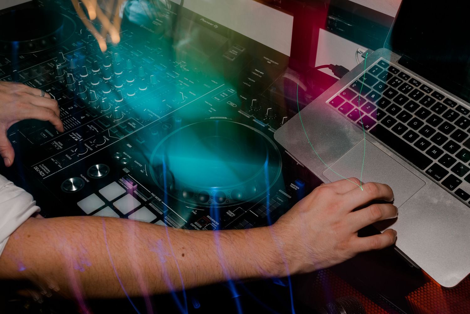 Hands type on laptop keyboards in dark room with vibrant blue and pink lighting creating a moody atmosphere.