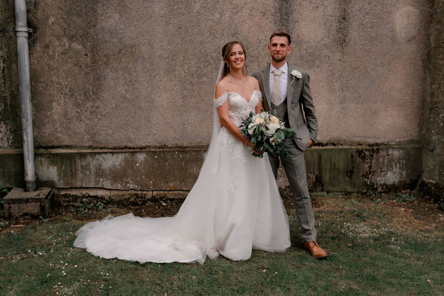 A bride and groom pose together outdoors against a textured wall, she in a flowing white gown and he in a gray suit.