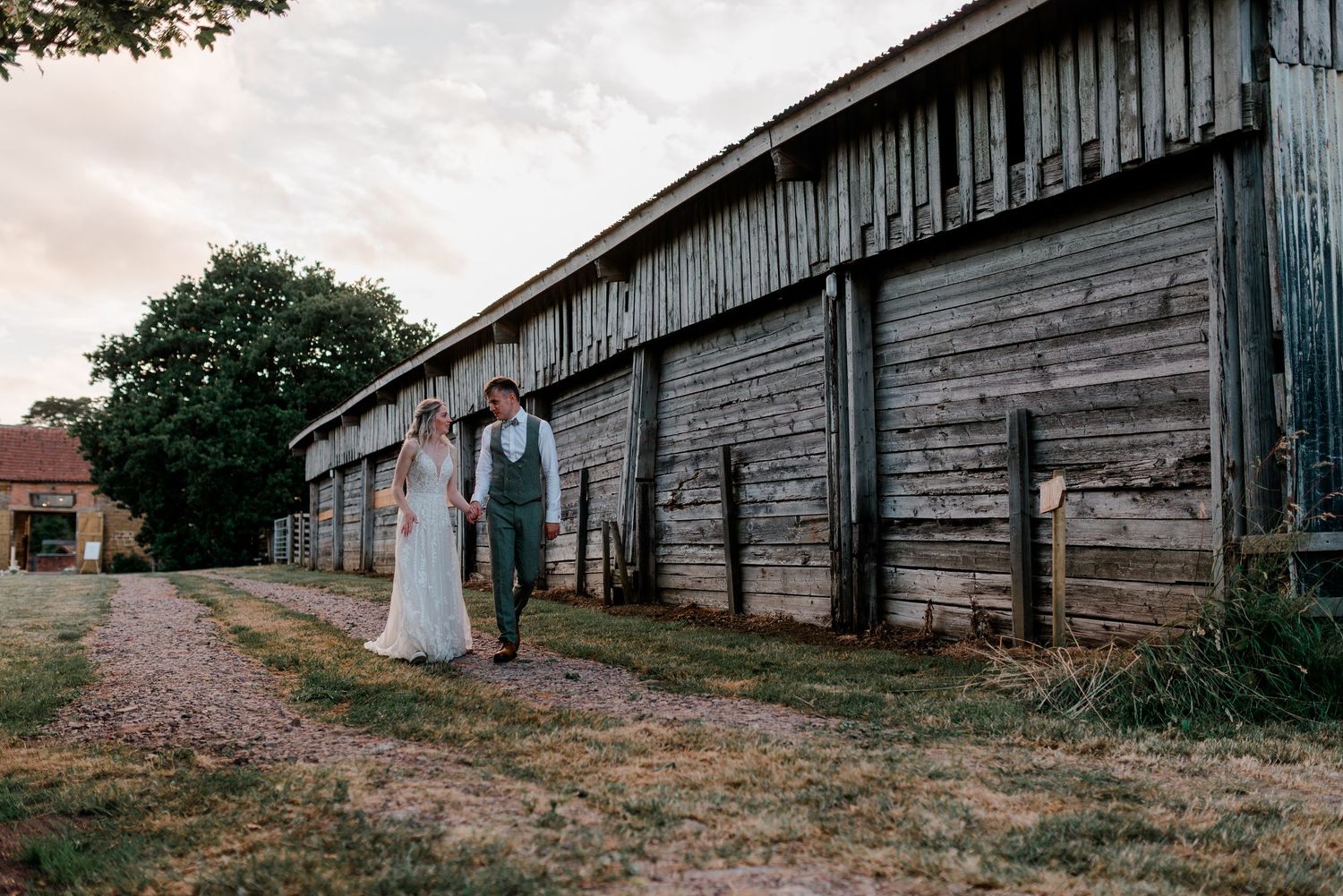 Newlywed couple standing beside weathered wooden barn on dirt path at sunset.