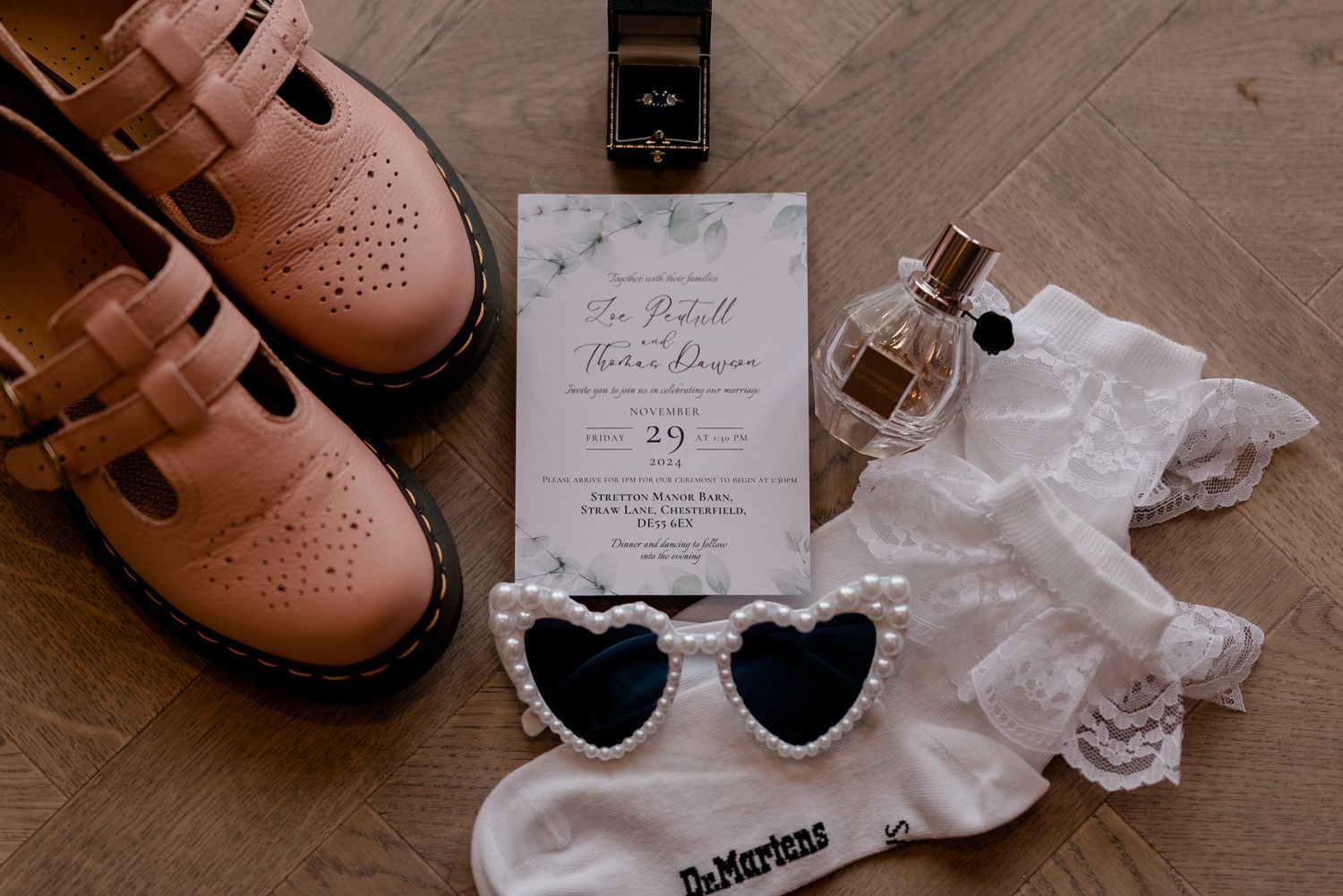 Wedding details flatlay with brown boots, heart sunglasses, lace socks and an invitation on wooden floor.