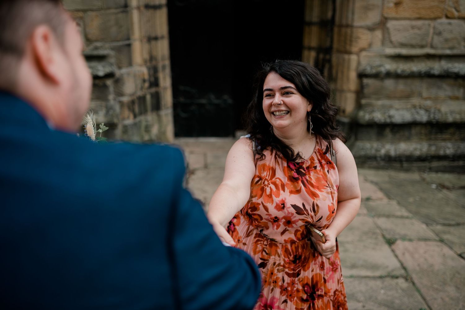 A person in a blue suit chats with someone wearing a vibrant orange floral dress against a stone building backdrop.