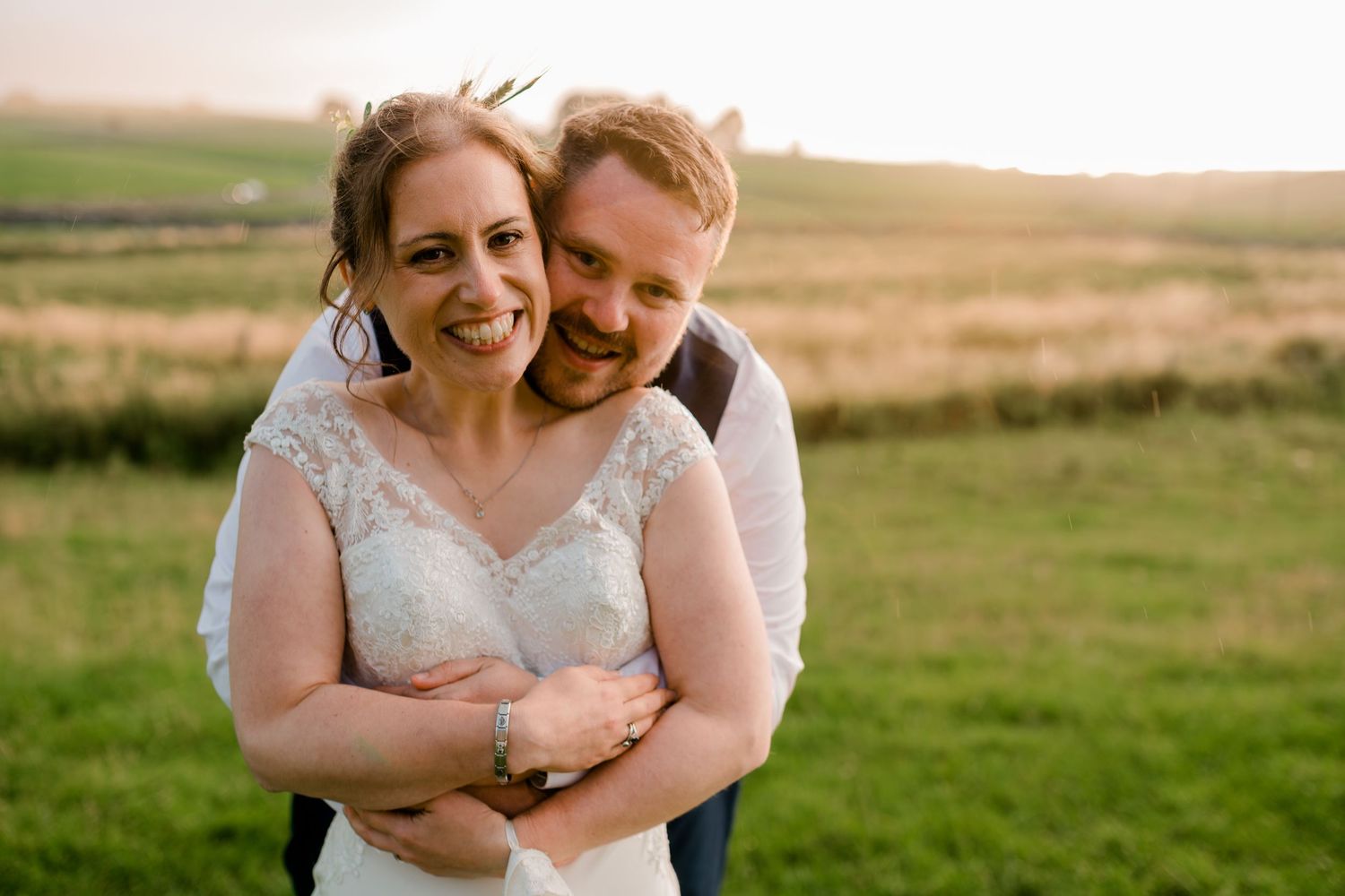 Smiling bride and groom embrace warmly in a sunlit green countryside field during golden hour.