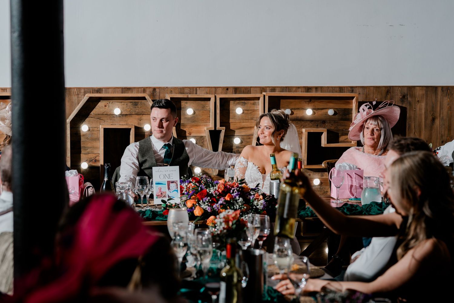 Bride and groom seated at decorated wedding reception table with colorful floral centerpiece.
