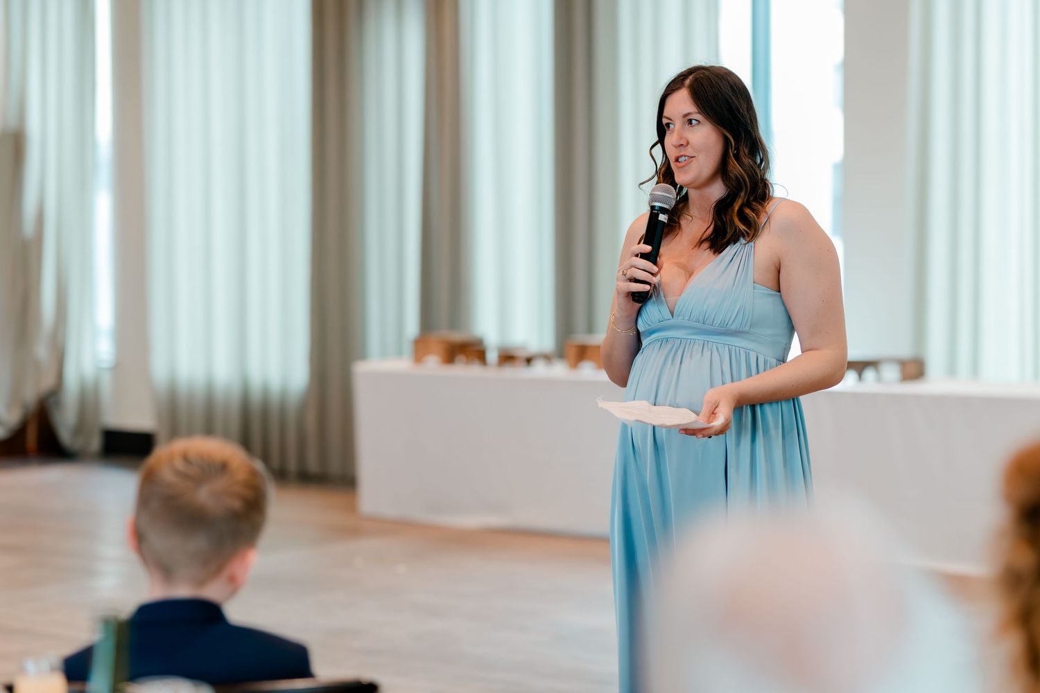 Woman in blue dress giving a speech at a wedding reception while holding a microphone and notes.