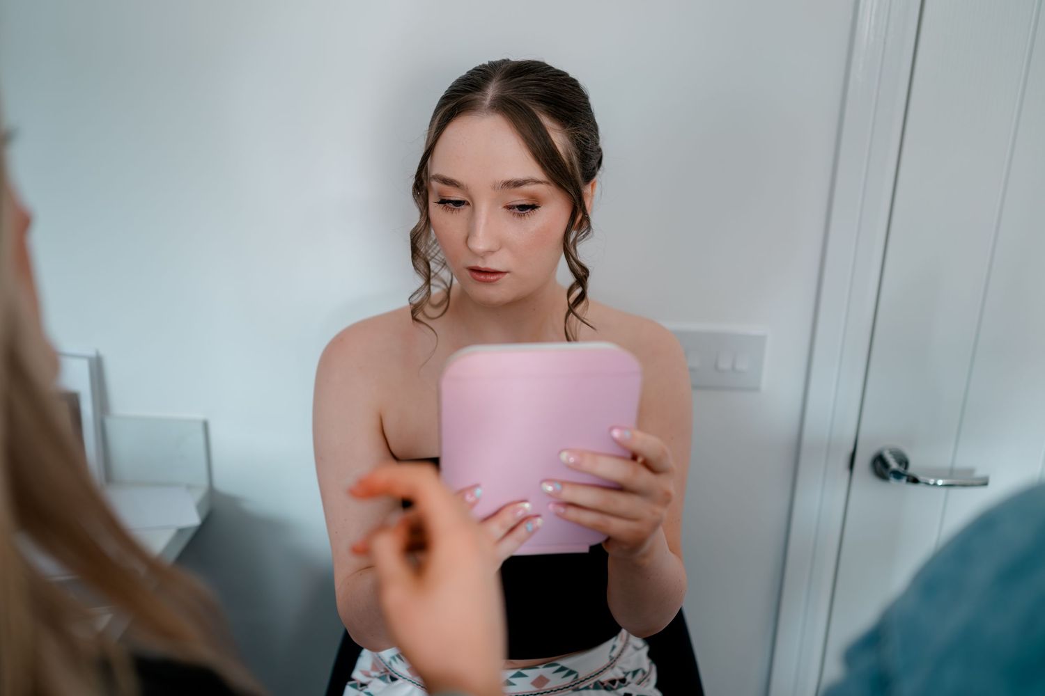 Young woman in formal attire examines a pink box while getting ready, reflected in a mirror.